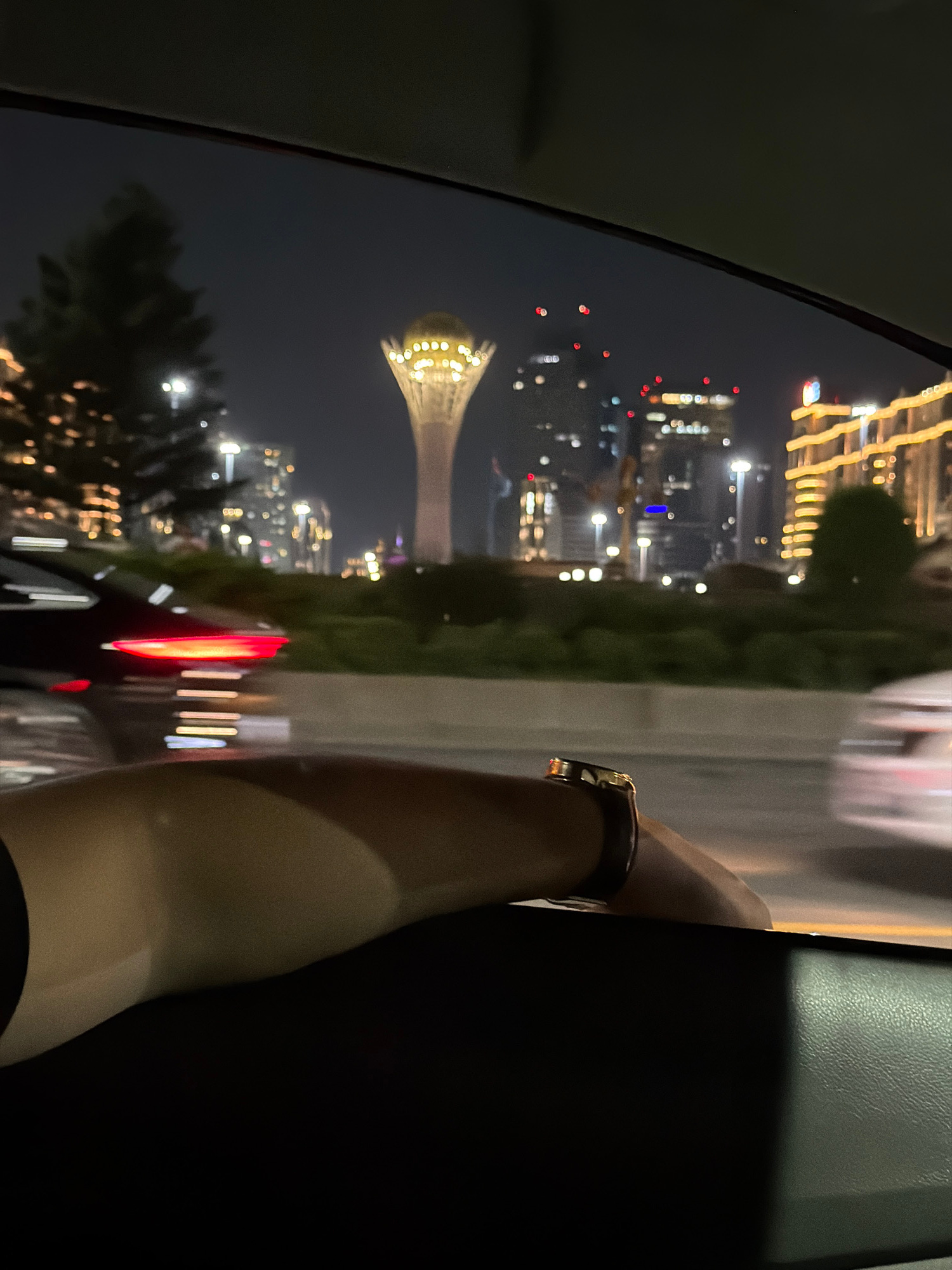 The image captures a night-time view from inside a vehicle, looking out through a window at a cityscape. In the foreground, a person's arm is visible, resting on the window sill, with a watch on the wrist. The arm and wrist are illuminated by ambient light, casting shadows.
The background reveals a blurred urban landscape at night. A prominent structure, appearing to be a tall, slender tower topped with a golden, illuminated sphere, stands out against the dark sky. This structure is likely the Bayterek Tower, a national monument in Astana, Kazakhstan. Surrounding the tower are other buildings with illuminated windows and facades, suggesting a well-lit urban center. The motion blur of passing vehicles is evident on the road in the mid-ground, indicated by streaks of red from taillights and white from headlights. Greenery, possibly trees and bushes, lines the side of the road, further blurring into the background due to the vehicle's movement. The overall atmosphere is one of nighttime travel through a modern city.