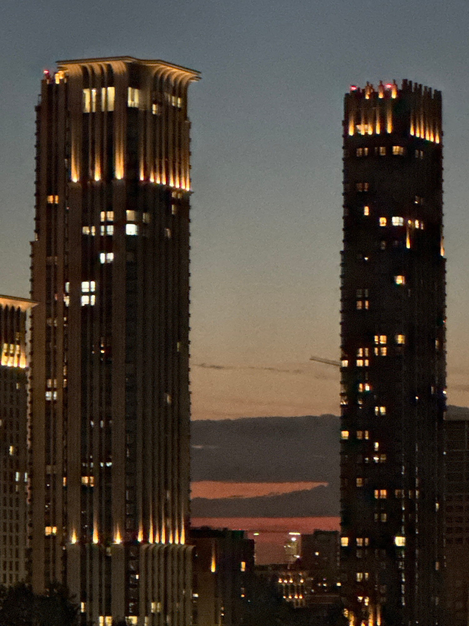 This media file presents an urban cityscape at night, likely viewed from a height in Moscow, Russia. Two prominent, tall modern high-rise buildings dominate the frame.
The building on the left side (when viewed upright, as the provided image is rotated) features a distinct, subtly curved architectural design at its upper sections. Horizontal ledges or balconies line its facade, reflecting warm light from numerous internal windows. These windows glow with scattered light, creating a mosaic of bright rectangles against the building's dark exterior.
The building on the right is similar in height and contemporary design, also exhibiting a dark facade punctuated by numerous illuminated windows. Its upper portion also shows architectural details that catch and reflect light.
Between and behind these two primary structures, the dark sky transitions to a softer, warmer glow near the horizon, displaying subtle orange and reddish hues, indicative of a lingering sunset or the distant luminescence of city lights. Other less defined buildings with faint lights are visible further in the background.
The scene captures the quiet, illuminated presence of urban architecture after dusk. No people or specific activities are visible, and there is no discernible text. The image conveys a calm, observational perspective of a metropolitan area at night.