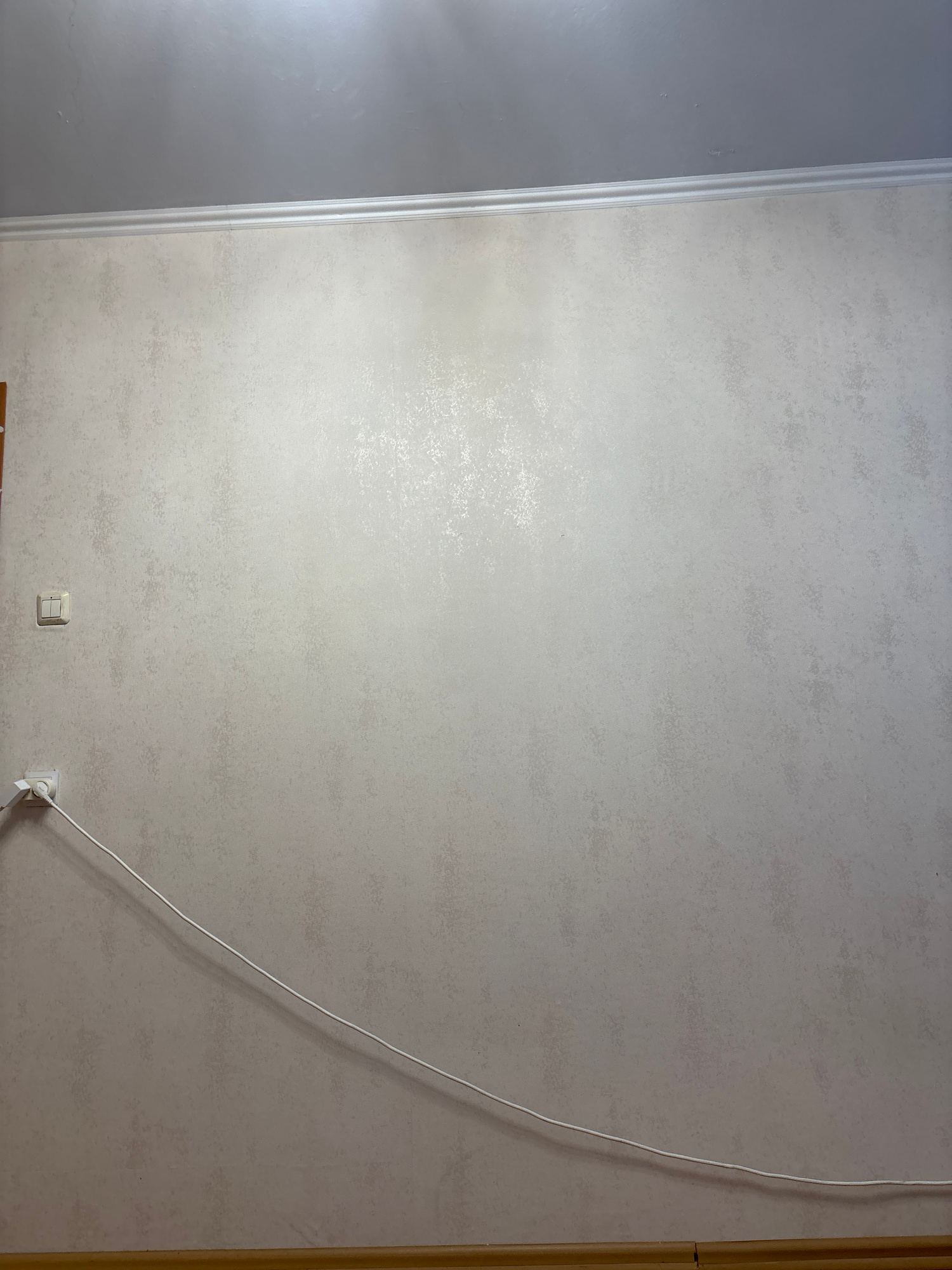 The image shows an interior wall with textured wallpaper in a light, neutral color. A white electrical cord runs diagonally across the lower portion of the wall, connecting to an electrical outlet and a switch plate. The ceiling is visible at the top of the image, with a white decorative molding separating it from the wall. The floor is partially visible at the bottom, appearing to be a wooden baseboard. The scene depicts a domestic or office interior. The image does not contain any information that would allow for the determination of its location in Saint Petersburg, Russia.