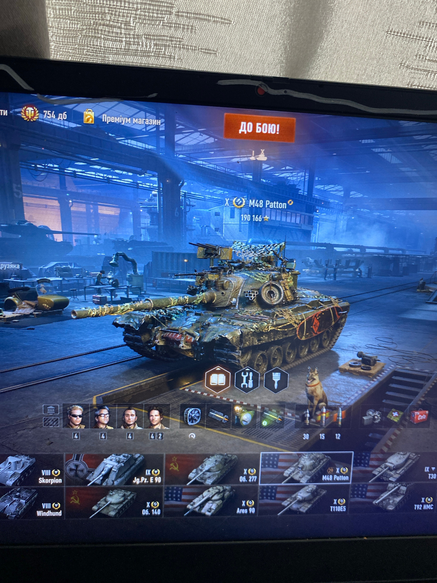 The image is a screenshot of a computer game interface, likely a tank simulation game like World of Tanks. The main subject is a detailed 3D model of a damaged tank in a dimly lit, industrial-looking hangar. The background shows more tanks and machinery.
The user interface elements visible include:
- A currency or resource counter showing "754 дб" (likely Russian for currency units).
- A "Premium Shop" icon and text.
- A large orange button with the text "ДО БОЮ!" (Russian for "To Battle!").
- Information about the currently selected tank: "M48 Patton" with a tier indication of "190 166".
- A row of crew portraits with associated numbers, possibly representing crew skill levels.
- A selection of other tank icons at the bottom of the screen, each with a tier, name, and country flag (e.g., "VIII Skorplon", "IX Jg.Pz. E 90", "X 06. 140", "X M48 Patton", "IX T30", "VIII Windhund", "X Ares 90", "X T110E5", "X T92 HMC").
- Various icons that could represent game functions like customization, crew management, or consumables.
- A dog statue is positioned near the tank.
There is no information in the image that indicates the location is in the city of Szczecin, Poland. The environment depicted is a virtual, in-game hangar.