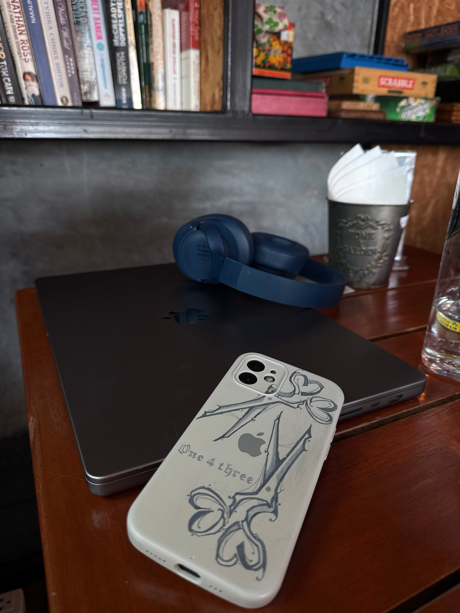 On a wooden table, a silver laptop is positioned to the left, with a pair of blue JBL headphones resting on top. To the right of the laptop, a white iPhone with a decorative case featuring an apple logo and the text "One 4 three" is placed. Further to the right, a glass of water with a yellow label is visible. In the background, a shelf holds various books and a tin container with white paper products. The scene appears to be indoors, possibly in a cafe or a home, in Rawai, Thailand.