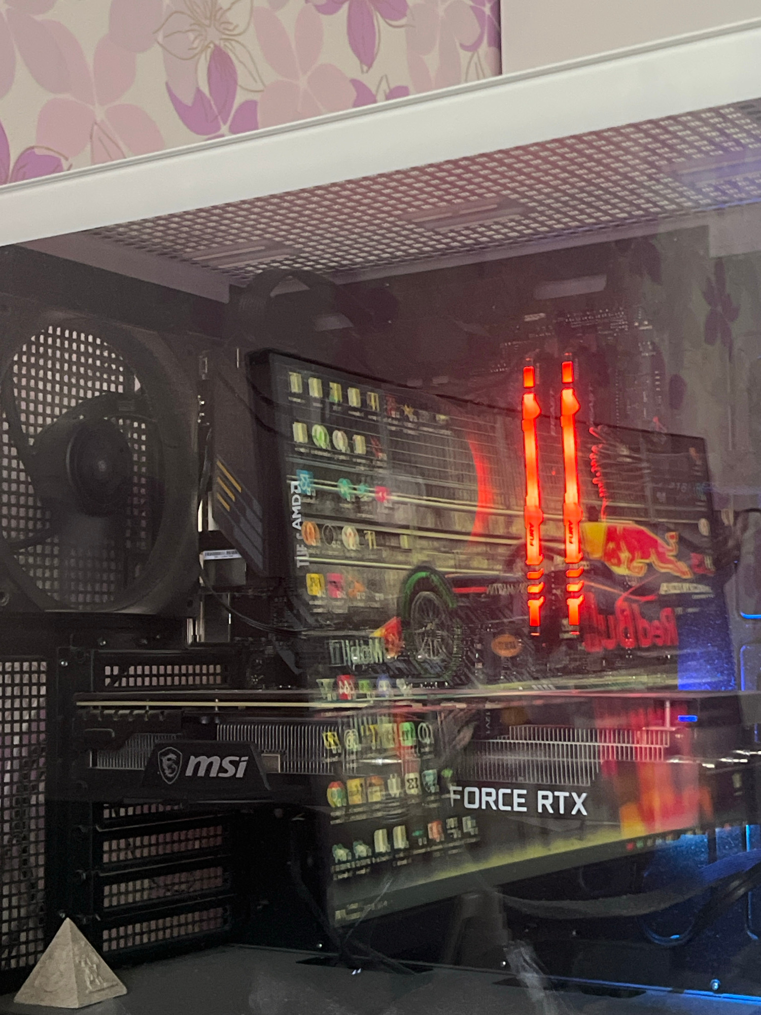 The image shows the interior of a computer case. Visible components include a large fan on the left, a graphics card with "FORCE RTX" branding, and RAM modules that are lit with red light. The graphics card has a design featuring a race car and the Red Bull logo. The computer is situated indoors, with a floral-patterned wallpaper visible in the background. The specific location in Kostroma, Russia, cannot be determined from the image provided.