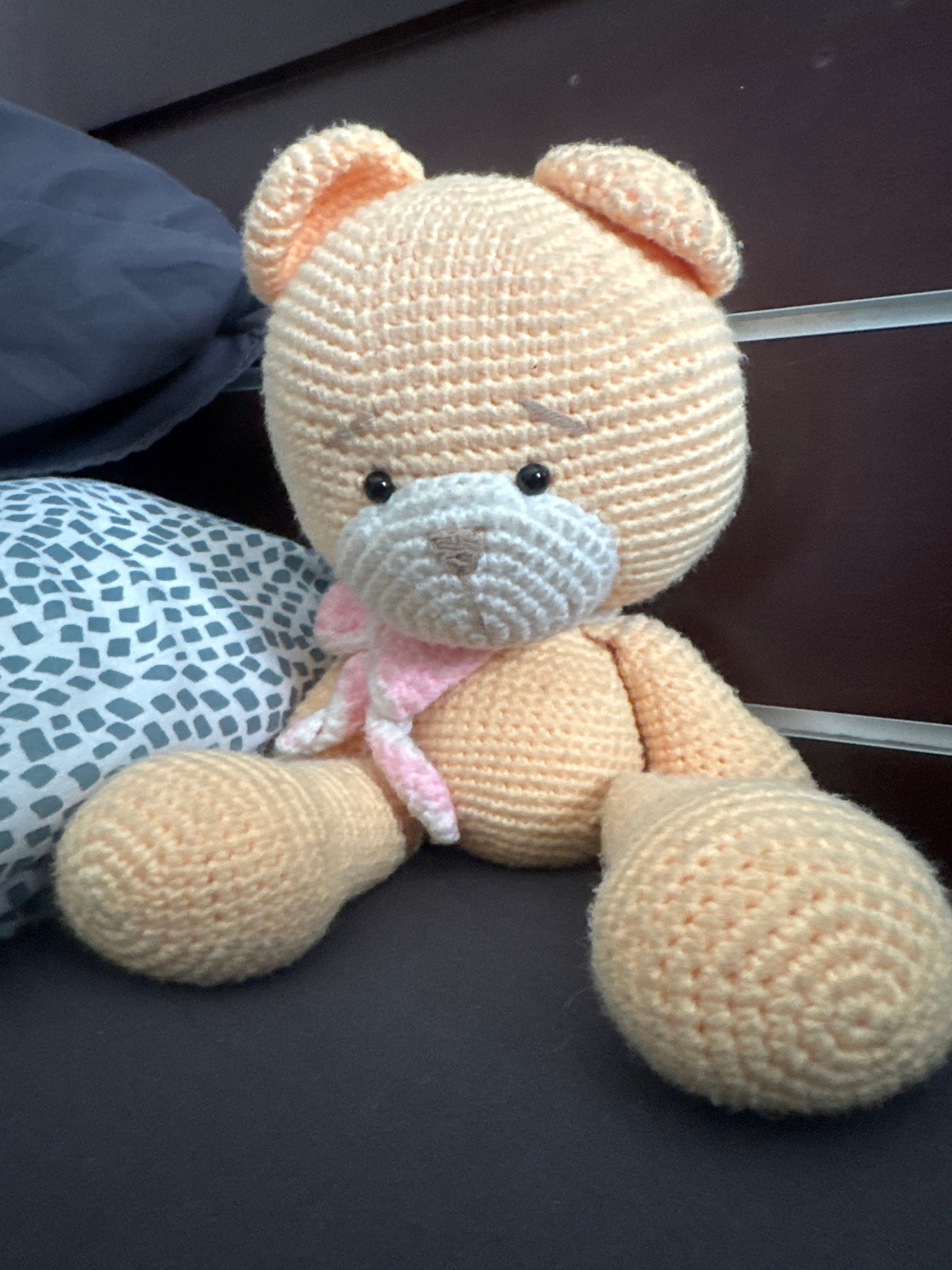 A crocheted stuffed bear with peach-colored fur sits on a dark surface, positioned in the center of the frame. The bear has black bead eyes, a white muzzle, and a pink and white striped scarf around its neck. Its ears are small and rounded, and its arms and legs are short and stout. To the left of the bear is a pillow with a gray and white speckled pattern, and behind the bear is a dark, textured surface, possibly furniture. The lighting is soft and appears to be coming from the right. This scene is located in a bedroom in Kuta, Indonesia.