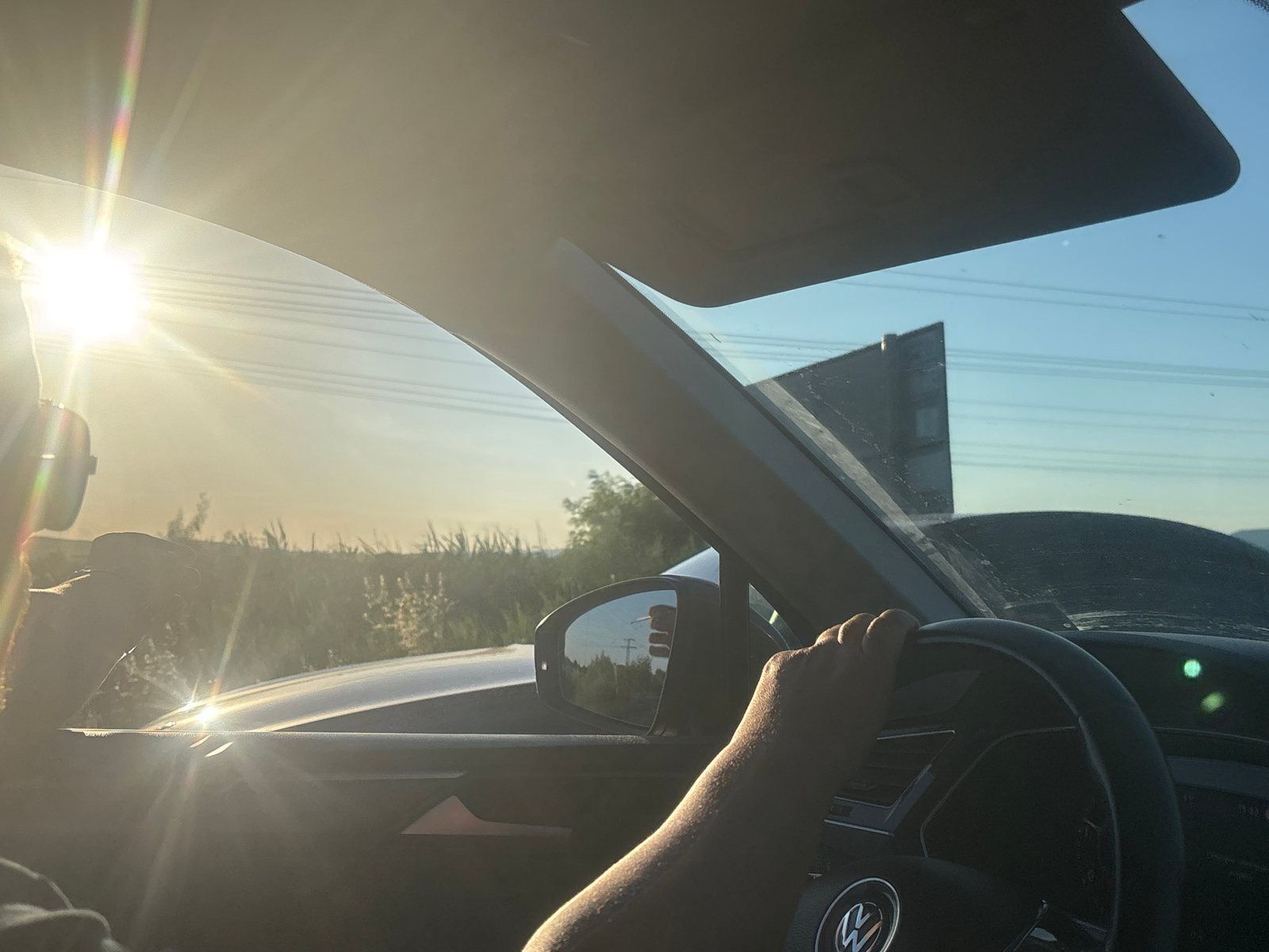 The image captures a view from inside a car, looking towards the right and slightly ahead. A person's right hand is visible, gripping the steering wheel of the car. The sun is intensely bright, appearing on the left side of the frame and casting strong lens flares and glare across the windshield and the interior of the car. The sky is clear and blue. Outside the car, a landscape of vegetation, possibly fields or tall grass, is visible under the bright sunlight. In the rearview mirror, a reflection shows trees and a telephone pole. The dashboard of the car is partially visible, with a logo that appears to be from Volkswagen. There are no discernible landmarks that would specifically indicate the city of Nesebar, Bulgaria, or any other specific urban location. The overall impression is of a sunny day, possibly during sunrise or sunset given the low angle of the sun and the intensity of the light.