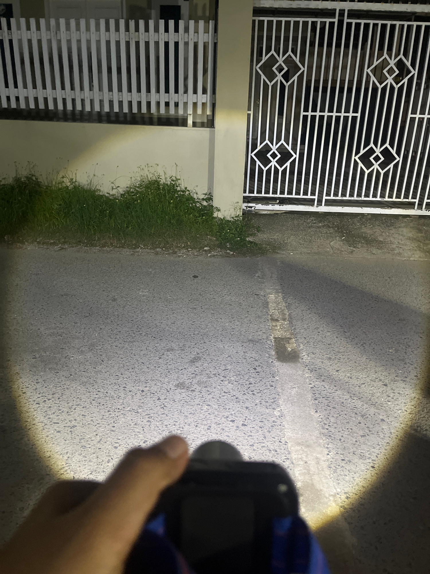 The image depicts a close-up view of an asphalt road at night, illuminated by a bright flashlight beam. The flashlight, held by a person whose hand is partially visible in the foreground, casts a strong light on the road surface, revealing its textured composition. A white line, likely a road marking, is clearly visible running horizontally across the illuminated area.
To the left of the road, a white metal fence with a geometric diamond pattern is visible, suggesting the edge of a property or building. Adjacent to the fence, a strip of green grass grows, indicating an outdoor setting. The background is dark, consistent with nighttime. The presence of the flashlight and the focus on the road suggest that the person is likely examining the road surface or using the light for navigation or a specific purpose.
The overall scene is dark and possibly damp, given the reflection of light on the road. The image was taken in Lhokseumawe, Indonesia, suggesting a tropical or subtropical environment. There is no visible text in the image.