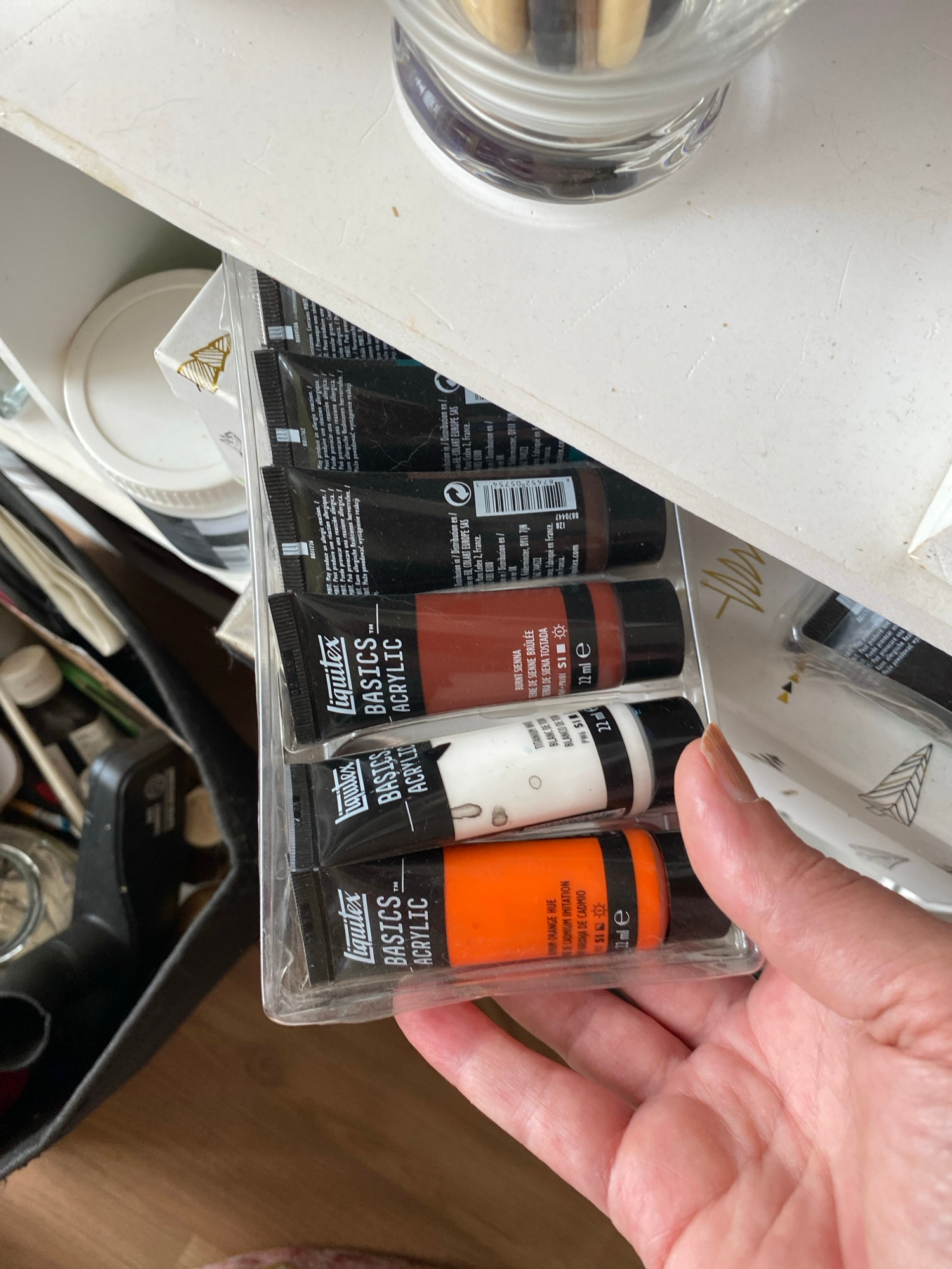 The image displays a close-up view of a hand holding a clear plastic package containing four tubes of acrylic paint. The scene appears to be an indoor setting, likely a crafting area or art studio, in Bílina, Czechia.
The subjects include a person's hand, which is lightly tanned with short, natural nails, grasping the paint set from above. The main objects are four tubes of "Liquitex BASICS ACRYLIC" paint, neatly arranged within a transparent plastic blister pack. From right to left, the colors clearly visible are a bright orange, white, and a reddish-brown. The leftmost tube is dark brown or black, with its label partially obscured.
Notable details include the brand "Liquitex BASICS ACRYLIC" prominently displayed on each tube. Each tube specifies a volume of "22 ml e." The visible paint colors are "CADMIUM ORANGE HUE" (also labeled "NARANJA DE CADMIO"), "TITANIUM WHITE" (also "BLANC DE TITANE"), and "BURNT SIENNA" (also "TERRE DE SIENNE BRÛLÉE" and "TIERRA DE SIENA TOSTADA"). Product codes like "PW6 SI" and "PSI" are also present.
The packaging contains warnings about potential allergic reactions in multiple languages, including English ("May produce an allergic reaction"), French, Spanish, Italian, German, and Polish. Distribution information indicates "COLART EUROPE SAS" with addresses in "Mans Cedex 2, France" and "Adderminster, DP11 7," and also states "Fabriqué en France." Barcodes and additional product numbers like "8011736" and "8011762" are also visible.
In the background, glimpses of other art supplies, such as a clear jar with brushes and various white containers, suggest an active creative space. The surface beneath the paint set is light-colored, possibly white, with subtle textures and lines. A dark bag or container with additional items is visible in the lower right portion of the frame.