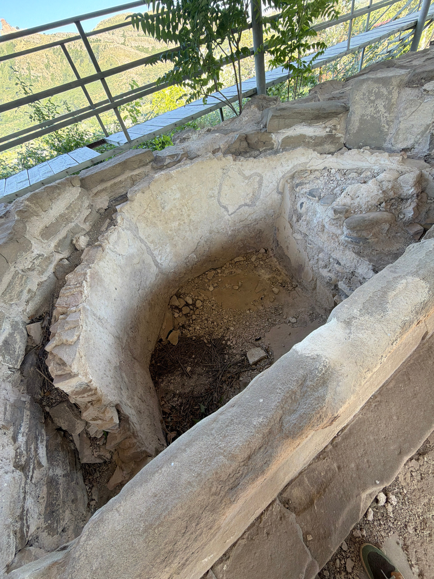 The image shows an outdoor archaeological site, likely in Mtskheta, Georgia, given the location context. The primary subjects are the remains of ancient stone structures, including a large, curved stone trough or basin. The stone appears weathered and aged, suggesting a historical setting. In the background, a metal fence or railing runs along the edge of the site, with lush green trees and foliage visible beyond it, hinting at a natural landscape or a preserved area. The overall impression is one of ancient ruins set within a natural environment, possibly a historical park or excavation site. The lighting suggests it is daytime, with clear, bright conditions. There is no visible text in the image.