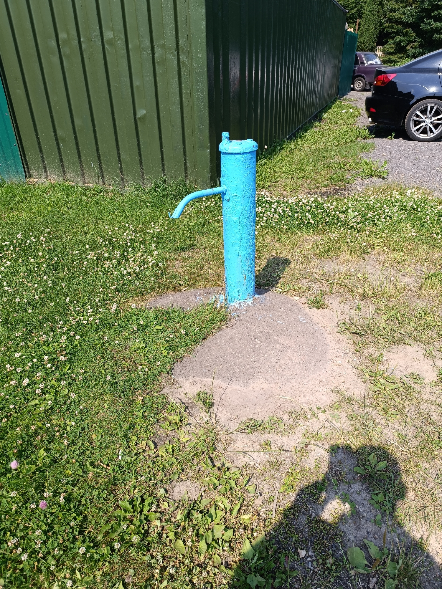 The image shows a blue manual water pump in front of a dark green corrugated metal wall. The pump is located on a patch of concrete surrounded by grass and clover. The wall appears to be part of a building or structure. In the background, a black car is parked on a gravel surface. The scene suggests an outdoor area, possibly a yard or public space. The lighting indicates it is daytime. There is a shadow of a person in the lower right corner of the image. The provided information specifies the location as Minsk, Belarus.