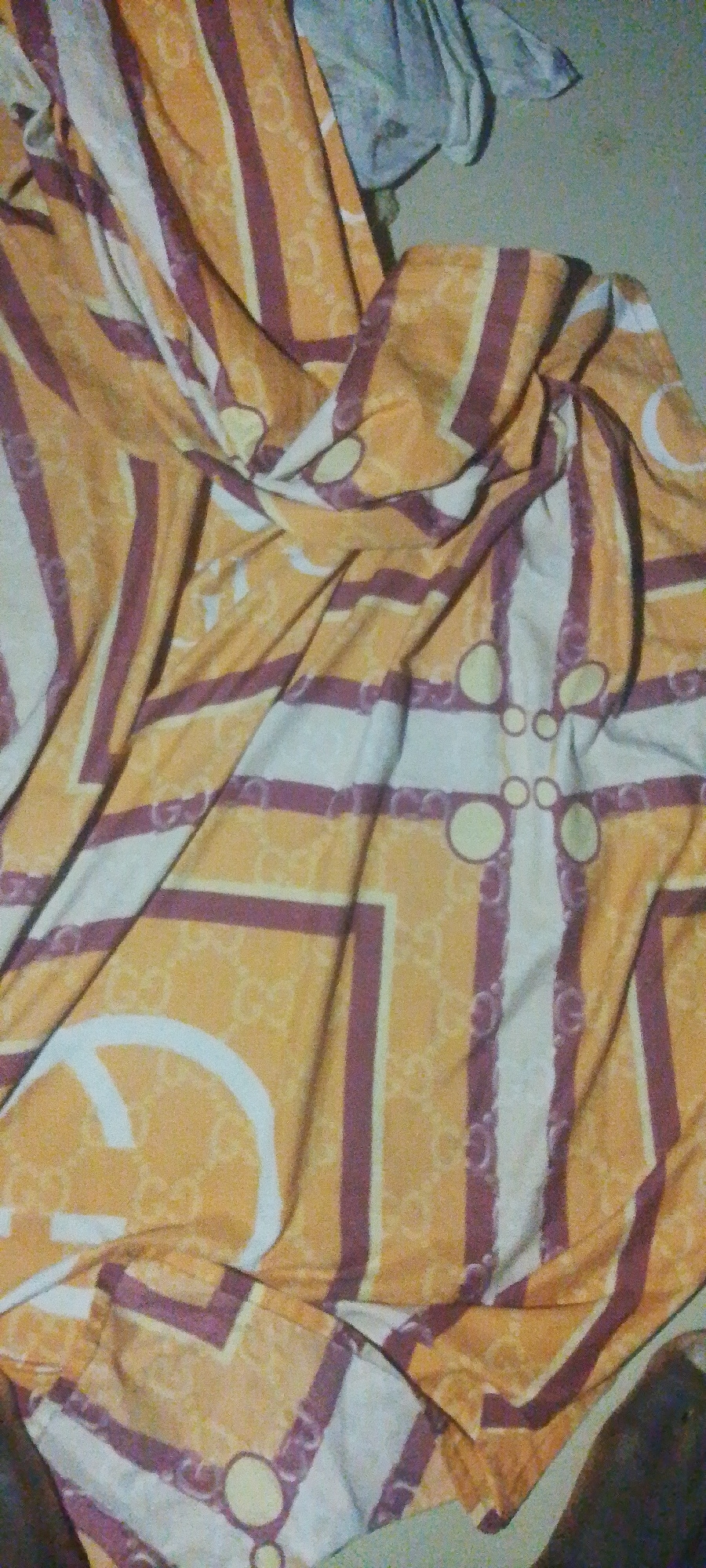 An overhead, slightly angled, medium shot captures a crumpled bedsheet with a bold pattern of orange, white, and purple stripes, and what appear to be repeating logos or symbols. The fabric is creased and folded, obscuring a clear view of the entire pattern. The background is a muted, textured wall, with some indistinct, lighter colored fabric draped in the upper left corner. The lighting is dim, suggesting an indoor environment. This scene is located in Jauro Yinu, Nigeria.
