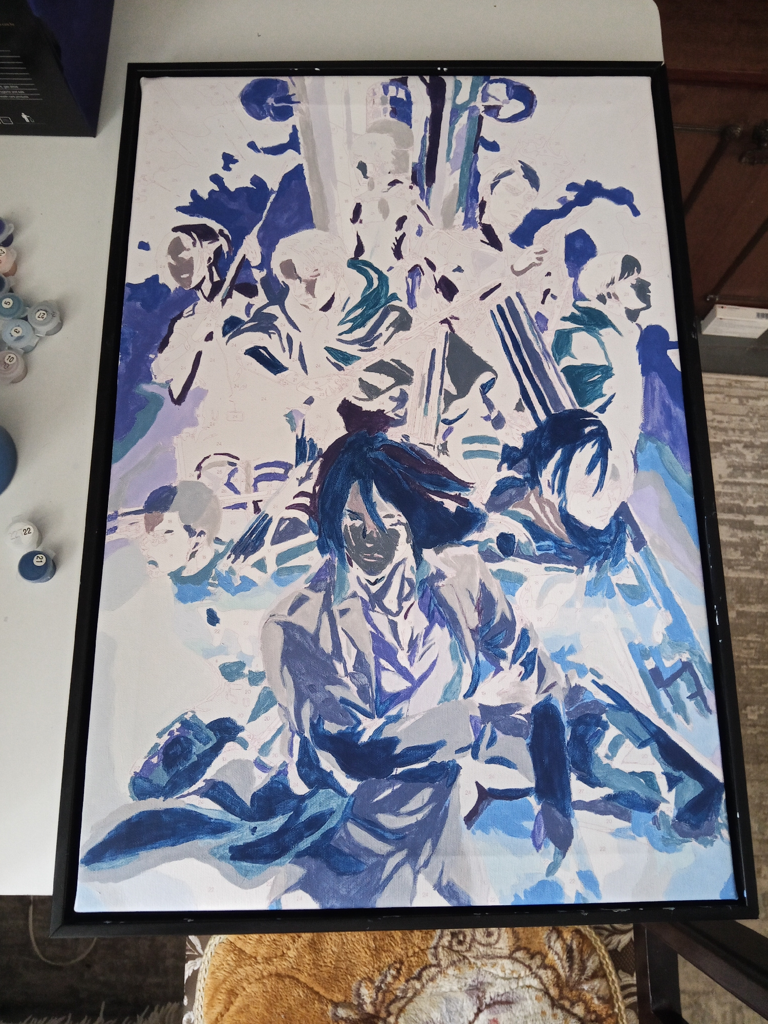 The image displays a partially completed paint-by-numbers canvas featuring a group of figures rendered in shades of blue and white. The canvas is framed in black and rests on a surface. In the foreground, several small containers of paint, labeled with numbers, are visible. The overall scene suggests an artistic endeavor in progress. The location in Qaratal, Kazakhstan, is not depicted or implied by any elements within the image.