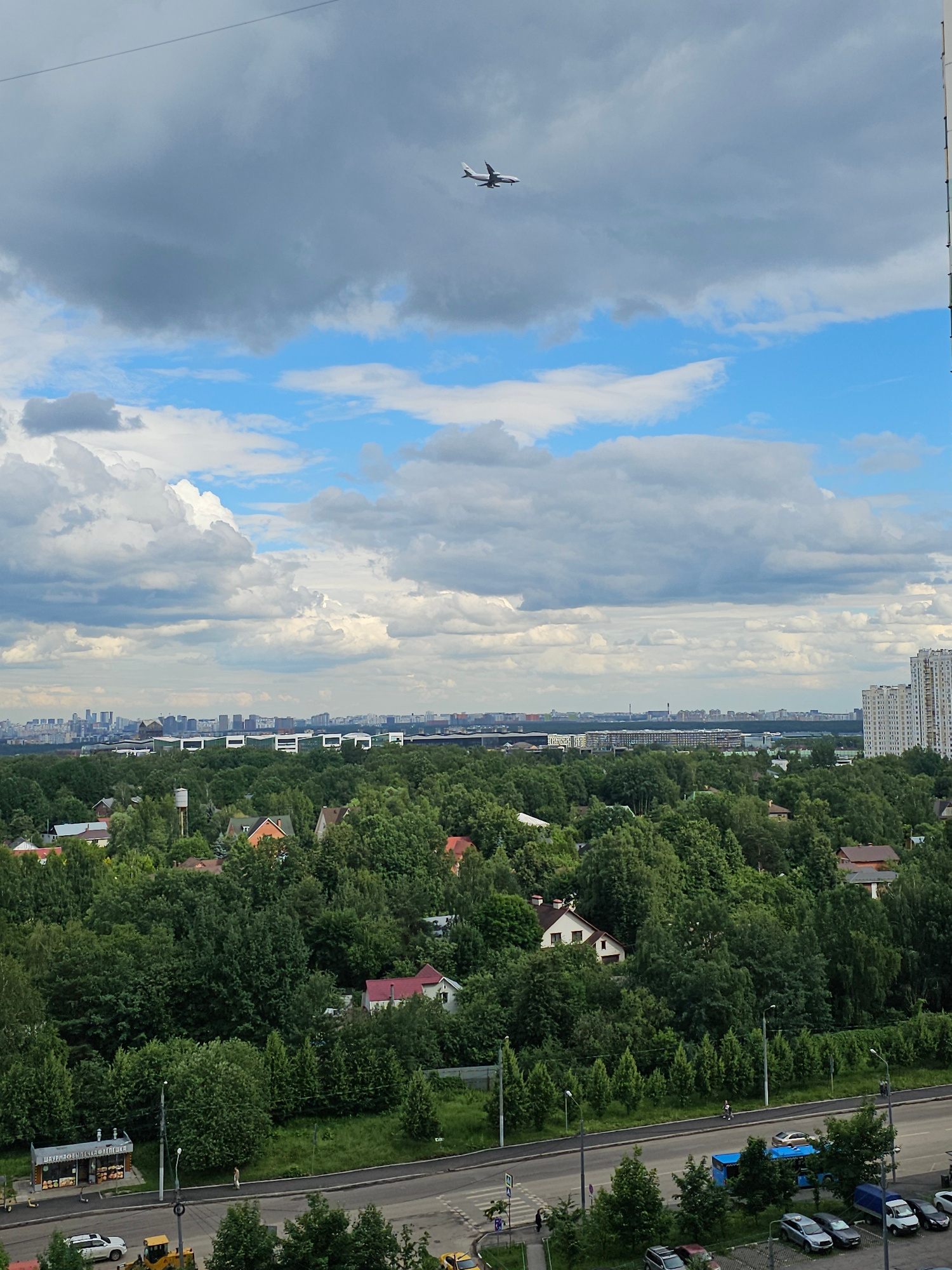 This is a high-angle, medium-distance, eye-level, vertical, color, daylight photograph taken from a building overlooking a suburban area of Odintsovo, Russia. The sky is partly cloudy with dramatic cumulus formations. An airplane is visible in the upper left quadrant, flying at a moderate altitude. Below the sky, a dense canopy of green trees stretches across the midground, punctuated by scattered residential houses with red and brown roofs. In the foreground, a paved road runs horizontally, with several vehicles including a white car, a yellow bulldozer, and a blue bus parked along its edge. A small commercial building with signage in Russian is situated on the left side of the road. The background features a distant cityscape with numerous high-rise buildings. The overall scene depicts a blend of nature and urban development.