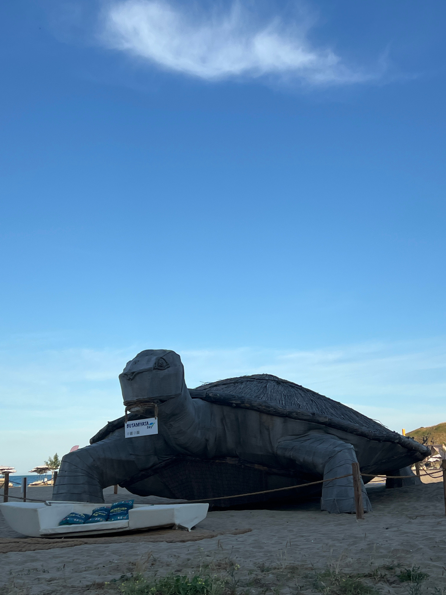 The image captures a large, dark grey, turtle-shaped sculpture situated on a sandy beach. The sculpture has a textured surface, resembling weathered stone or concrete, and its shell is thatched with straw-like material. A sign reading "BUTAMYATA bay" is affixed to the sculpture's neck. In front of the sculpture, a white, boat-shaped seating area is placed on the sand, furnished with four blue-patterned cushions. The background shows the sea and a clear blue sky with a few wispy clouds. The location is identified as Butamyata Beach in Sinemorets, Bulgaria, a popular tourist destination known for its unique rock formations and natural beauty.