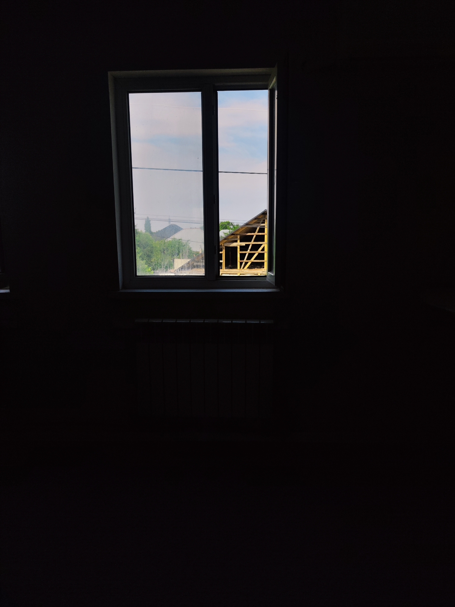 A rectangular, white-framed window is centrally located in a dark, interior room. The window is divided into two panes. Through the left pane, a cloudy sky and green trees are visible, along with power lines stretched horizontally across the frame. The right pane offers a view of a partially constructed building with a wooden frame, its roofline visible. Below the window, a dark-colored radiator with horizontal fins is positioned. The walls of the room are a dark, muted color, making the window the focal point of the image. The scene suggests an indoor perspective looking out onto a residential or semi-rural area. The location of Zhanadaur, Kazakhstan is not depicted or indicated in the image.