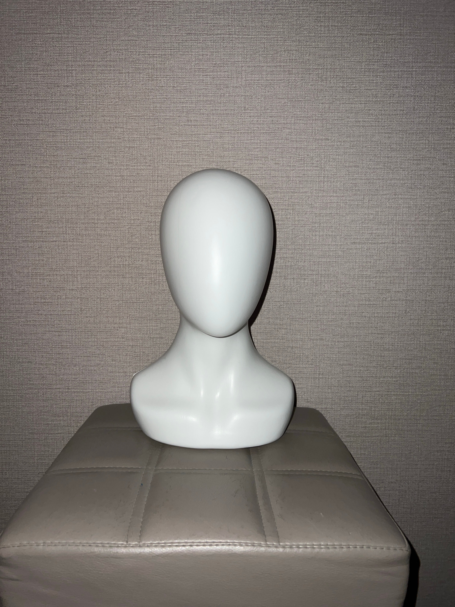 A white, faceless mannequin bust sits centered on a square, tufted, light brown ottoman. The ottoman is positioned in front of a textured, taupe-colored wall. The lighting casts a shadow to the left of the ottoman and mannequin. The overall scene is static and appears to be indoors.