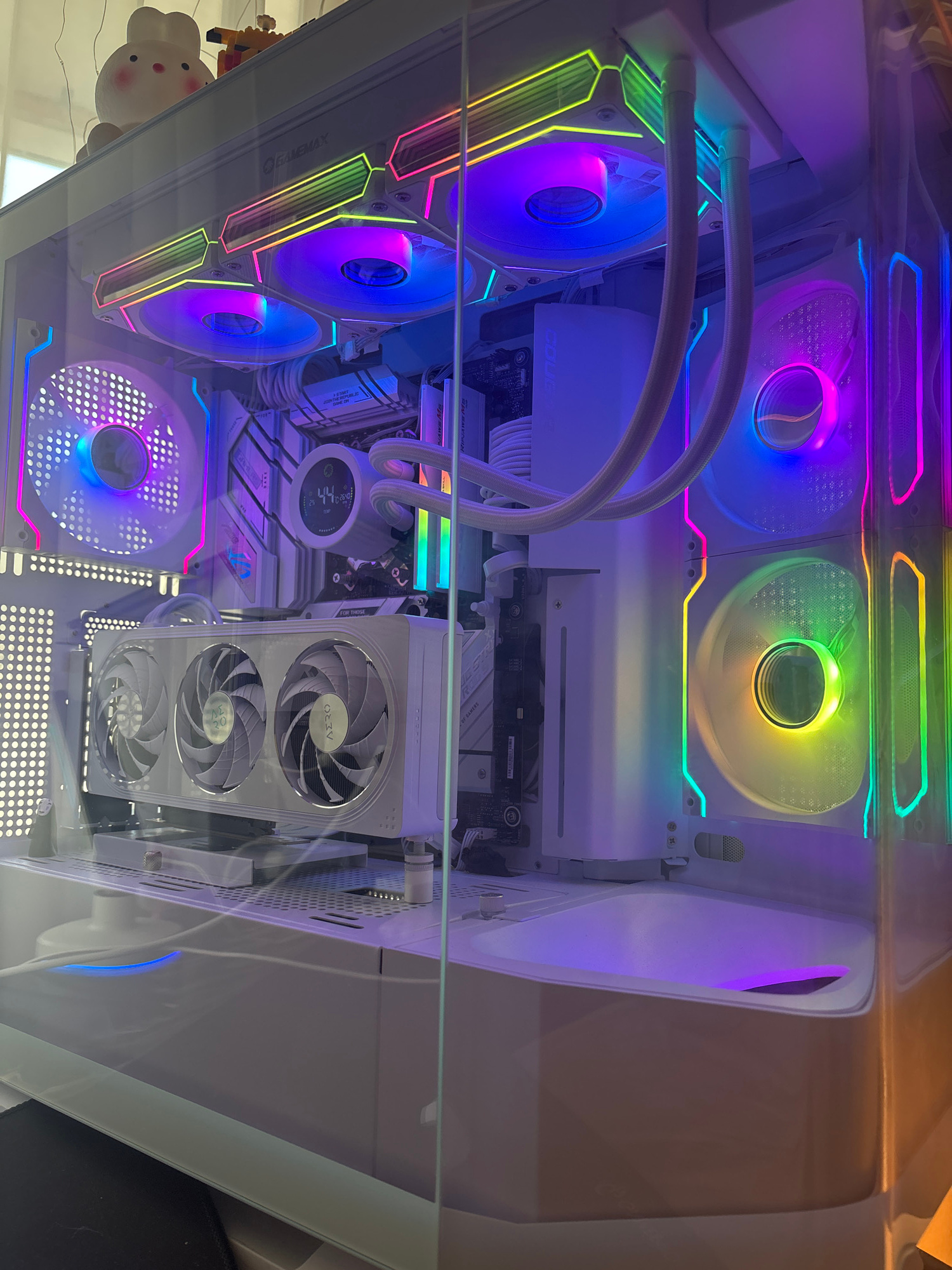 The image shows the interior of a white computer tower case, viewed from above through a glass panel. The computer components are illuminated by colorful LED lights, creating a vibrant and modern aesthetic.
Dominating the view are several large computer fans, some with ARGB lighting that cycles through a rainbow of colors, including blue, purple, pink, green, and yellow. Two prominent fans are positioned at the top of the case, while another set of three fans is mounted vertically on a component in the center-right. A smaller fan is visible at the bottom left, also illuminated.
Other visible components include a CPU cooler with a circular display, white braided cables, and what appears to be a graphics card or motherboard with heat sinks. The overall impression is that of a high-performance gaming or enthusiast PC, likely powered on and showcasing its lighting effects.
There is a small, white, plush toy with a round face and small ears on the left side of the image, near the bottom. There is also a hint of a yellow and black toy or object further to the left.
The lighting suggests the photo was taken in a dimly lit indoor environment, allowing the computer's LEDs to be the primary light source. No people are visible. The image is focused on the internal hardware of the computer.
Visible text includes "ASUS" on the motherboard, and branding on the fans that reads "DEEPCOOL" and "AZZA".