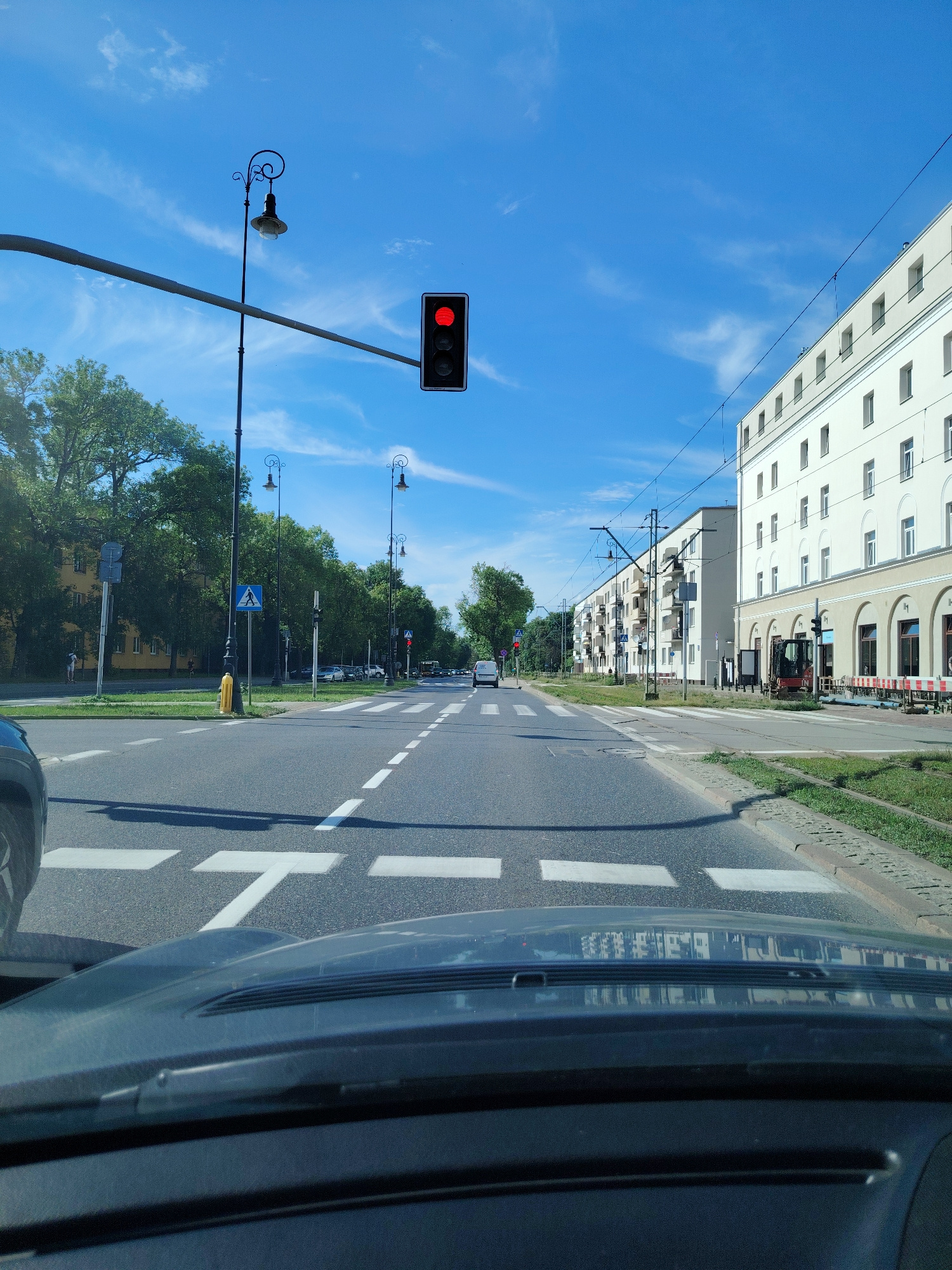 The image is a first-person perspective shot from inside a vehicle, looking forward through the windshield at a city street in Warsaw, Poland. The sky is bright blue with some wispy clouds. A red traffic light is visible overhead, indicating a stop. The street is paved and marked with white pedestrian crossing lines. On the right side of the street, there is a multi-story building with a light-colored facade and numerous windows. Some construction barriers and a small excavator are present in front of the building. On the left side of the street, there are trees, other buildings, and street lamps with decorative elements. There are cars and a tram visible further down the street, and a few pedestrians are seen on the sidewalks. The vehicle's dashboard is partially visible in the foreground. The scene appears to be a sunny day.