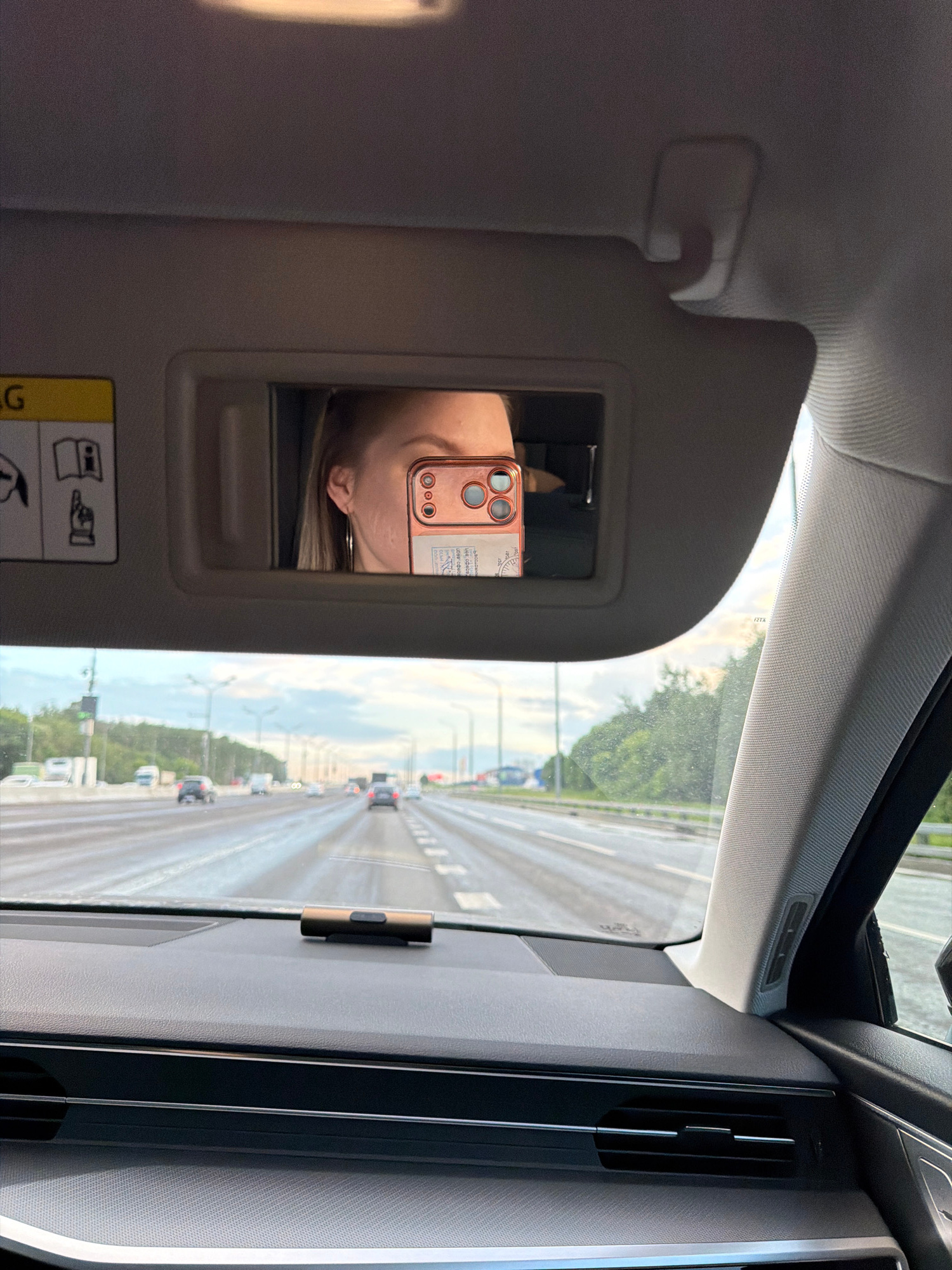 The image is a first-person perspective from inside a car, looking forward through the windshield towards a multi-lane highway. In the foreground, the dashboard is visible, with a small cylindrical object placed on it. Above the windshield, a sun visor is flipped down, revealing a mirror. Reflected in the mirror is the face of a woman, holding a rose-gold colored smartphone up to take the photo. She is wearing a hoop earring. The highway stretches into the distance, with several cars driving in both directions under a cloudy sky. The location is identified as Moscow, Russia.