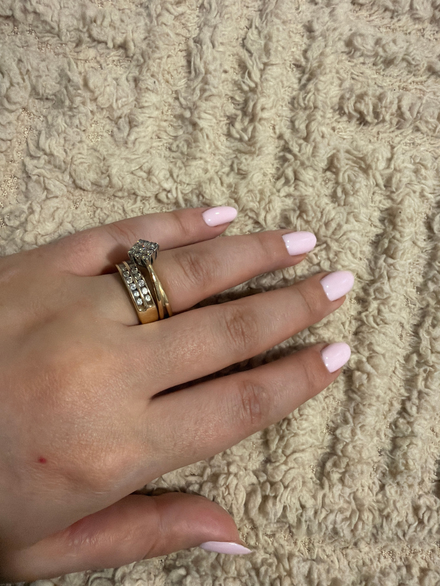 A close-up, eye-level shot shows a person's left hand with light pink nail polish resting on a textured, cream-colored fuzzy fabric. The hand is adorned with two gold rings. The top ring features a cluster of small, clear stones, possibly diamonds, arranged in a square shape. Below it, a second gold band is also set with rows of small, clear stones. The background is entirely filled with the fuzzy fabric. The image does not provide any information about the location in Yerevan, Armenia.