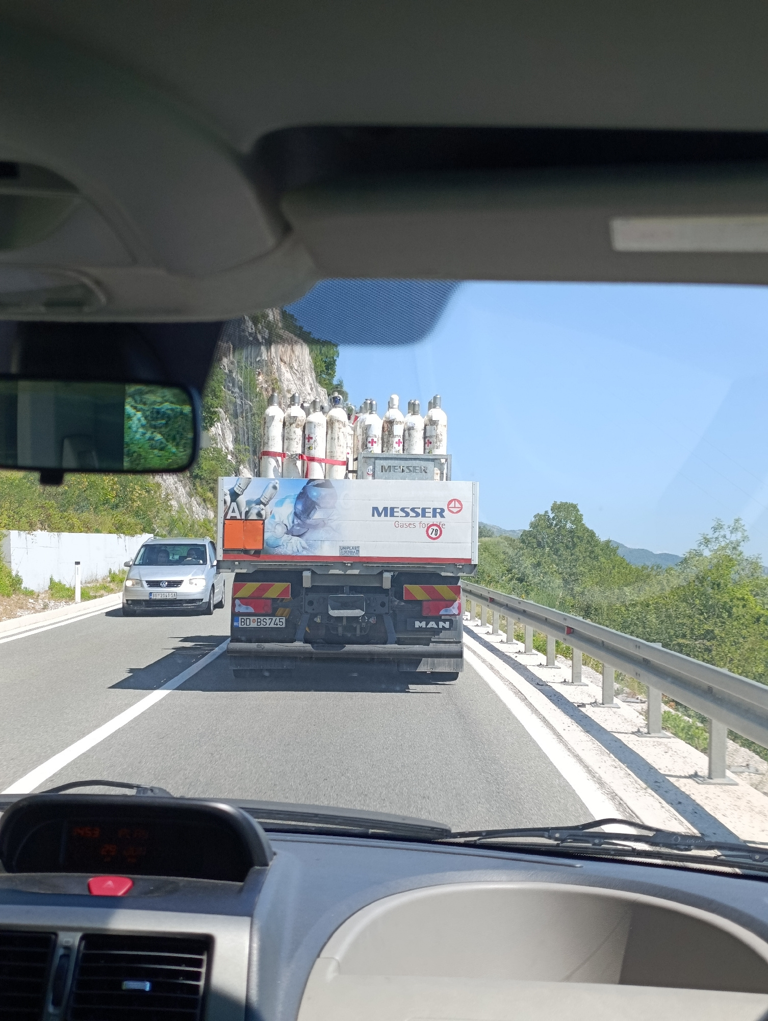 The image captures a view from inside a vehicle on a sunny day, looking out at a highway. A white truck with "MESSER Gases for life" branding and several large white cylinders is prominently in the foreground. The cylinders are strapped to the truck's flatbed and appear to be carrying industrial gases. The truck has "MAN" written on its rear. To the left of the truck, a silver Volkswagen hatchback is visible, with two occupants inside. The highway has a white dividing line and a metal guardrail on the right side. The surrounding environment includes lush green trees and a rocky, mountainous hillside. The sky is clear and blue. The interior of the vehicle shows the dashboard with an illuminated display and air vents.