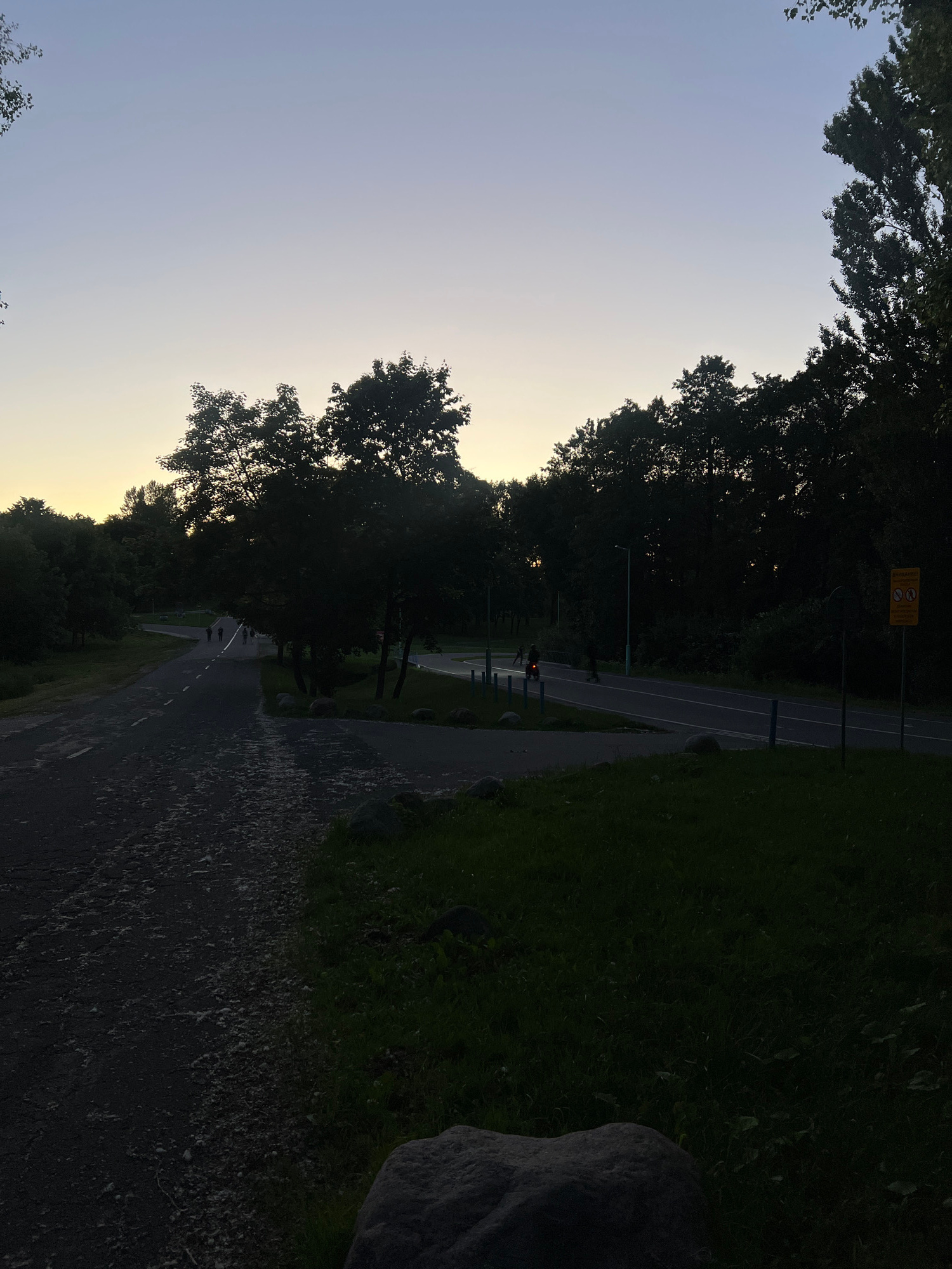 The image depicts a pathway in a park-like setting at dusk. A paved path curves to the left, and a rougher path leads to the foreground. Several figures are visible on the paved path in the distance, appearing to be walking or cycling. Trees line the sides of the path, forming a dense forest in the background. The sky is clear with a gradient from light blue at the top to a pale yellow near the horizon. A road with a few vehicles, including what appears to be a motorcycle with a rider, is visible to the right, bordered by trees and a grassy area. A yellow sign with symbols is posted on a pole to the right of the road. The overall impression is a tranquil, natural scene at the end of the day.