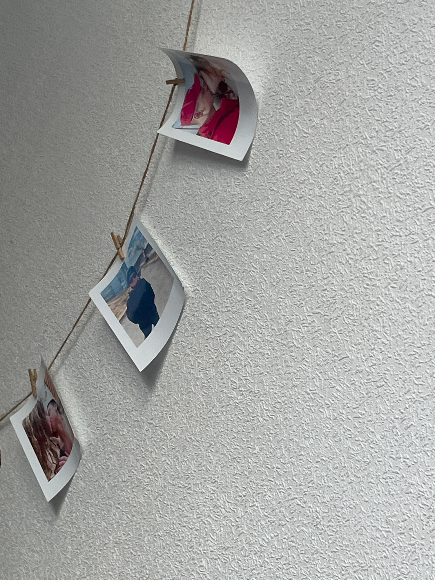 A thin string, likely twine, is strung diagonally across a textured, light-colored wall. Three photographs are clipped to the string with small wooden clothespins. The top photograph shows the face of a person wearing a bright pink garment. The middle photograph depicts a person in dark clothing, wearing a hat and sunglasses, standing in what appears to be an outdoor environment. The bottom photograph shows a close-up of two people, their faces are mostly obscured by their hair and the angle of the photo. The wall has a rough, stucco-like texture. There is no information available to suggest the location of this image is in Chelyabinsk, Russia.