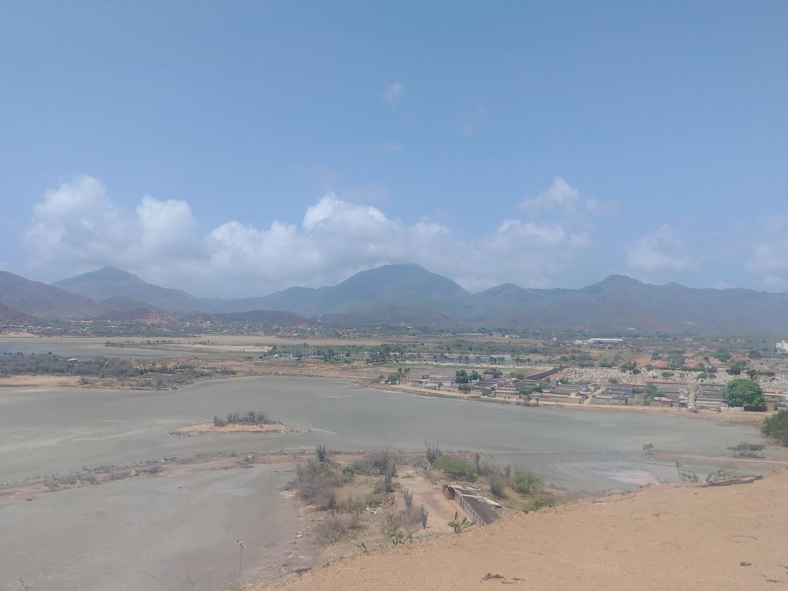 An elevated, wide-angle, eye-level view captures a panoramic landscape under a bright, partly cloudy sky. In the foreground, a dry, dusty, sandy slope descends into the frame. Beyond this, a shallow, grayish-brown body of water, possibly a salt flat or dried-up lake, dominates the mid-ground. Scattered across the water's edge and the adjacent dry land are sparse, arid vegetation including cacti and other scrub brush.
A small, rocky island with vegetation rises from the water in the mid-left. In the background, a town or city is nestled between the water and a range of hazy, mountainous terrain. Buildings with red-tiled roofs are visible, interspersed with trees and greenery. A large, rectangular structure, possibly an industrial building or a series of warehouses, is noted in the distance on the right side. The overall environment appears dry and sun-drenched, typical of a tropical or subtropical climate. The scene depicts the outskirts of Juan Griego, Venezuela, focusing on its coastal or near-coastal geography with elements of urban development encroaching on the natural landscape. No people or specific actions are discernible in the image.