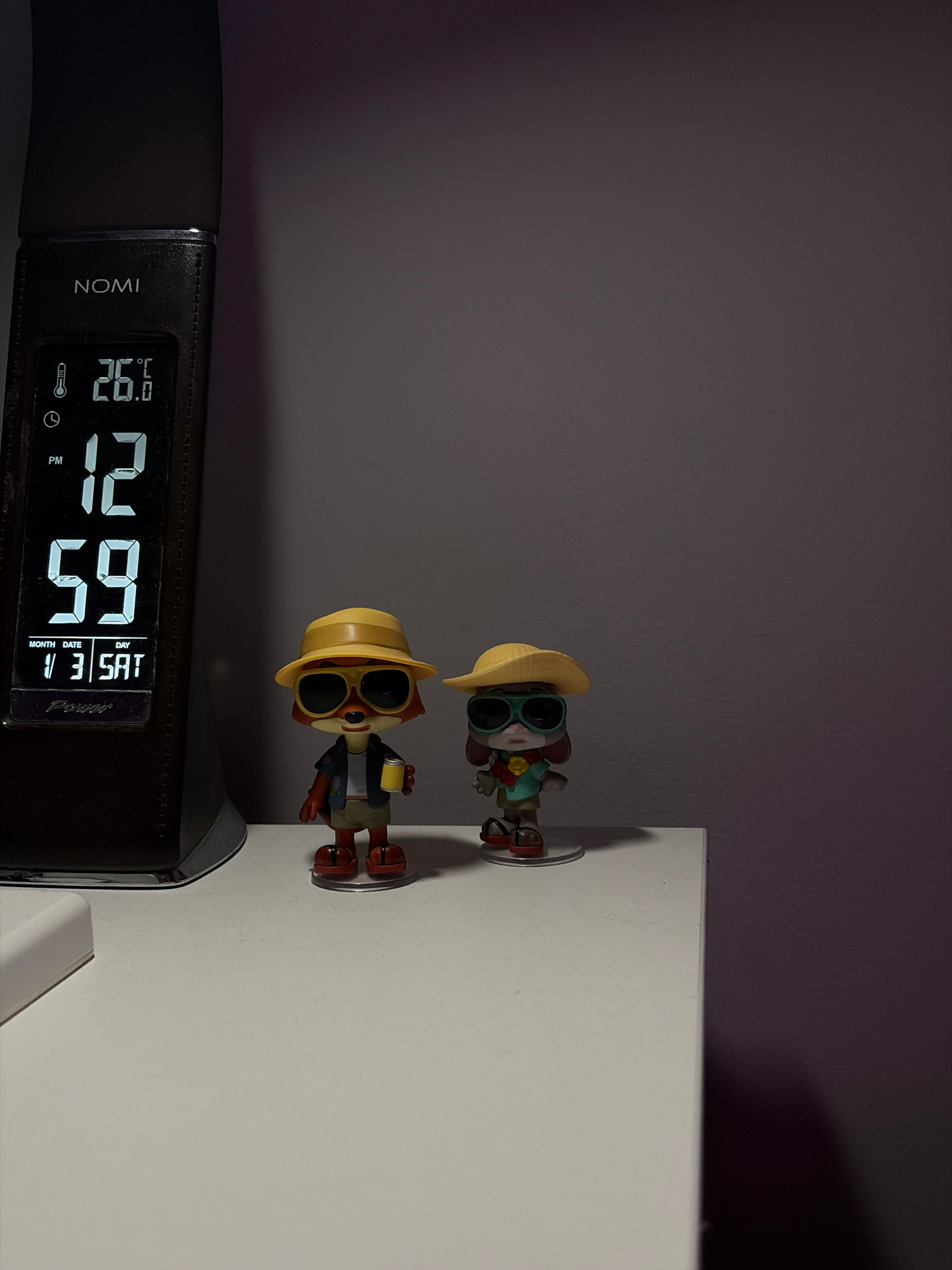 An indoor, eye-level, medium shot captures a bedside table with a digital clock and two small figurines. The clock, from the brand NOMI, displays the temperature as 26.6 degrees Celsius, the time as 12:59 PM, and the date as Saturday, January 3rd. To the right of the clock, two cartoonish figurines are positioned on the table. The first figurine, on the left, appears to be a red fox wearing a yellow hat, sunglasses, and a Hawaiian shirt, holding a yellow cup. The second figurine, on the right, is a rabbit wearing a yellow hat, large sunglasses, a green shirt, and brown shorts. The background is a plain, dark purple wall. The image is taken in Warsaw, Poland.