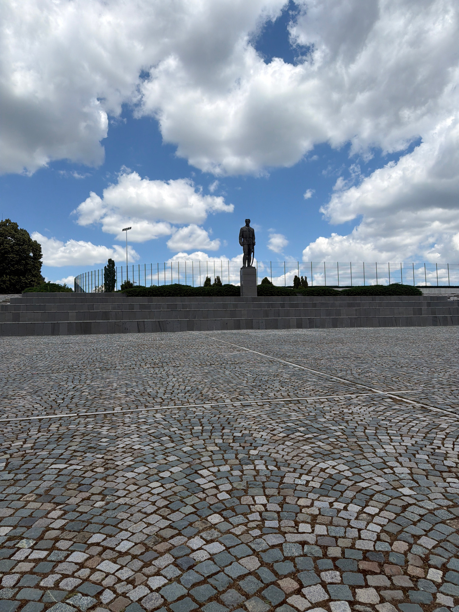 A full, eye-level, outdoor, slightly low-angle medium shot captures a cobblestone plaza in front of a tiered, stone amphitheater with a statue of a man on a pedestal at its center. The sky above is a mix of bright blue and dramatic, white cumulus clouds. The statue of the man, dressed in a military uniform and holding a sword, faces to the right, looking out over the plaza. Behind the statue, a short, manicured hedge runs along the top of the amphitheater, bordered by a fence with tall, thin posts. A tall, modern light fixture is visible to the left of the hedge, beyond the statue. The cobblestone ground in the foreground is laid out in a circular pattern, with lighter-colored stone lines marking sections of the plaza. To the far left, the dark green leaves of a large tree are visible against the sky.