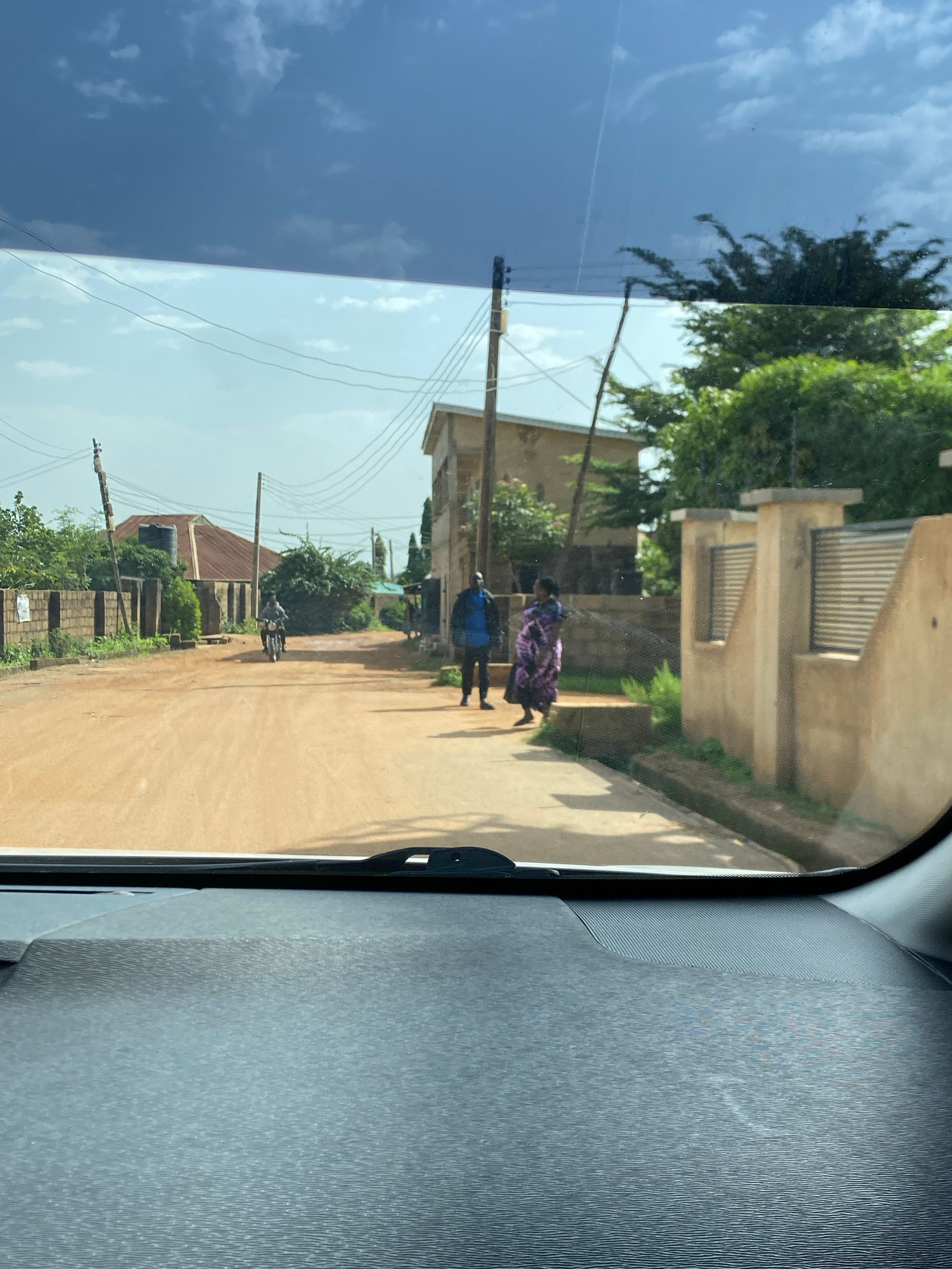 This image was taken from inside a vehicle, looking out through the windshield at a street scene in Bar Jirgi Summa, Nigeria. The road is unpaved and appears to be made of dirt or gravel. On the left side of the road, there is a brick wall and some vegetation. Further down the street, a motorcycle is visible with a rider. On the right side of the road, there are buildings with some architectural features such as metal gates and textured walls. Two individuals are standing on the right side of the road; one is wearing a blue shirt and black pants, and the other is wearing a patterned dress. The sky above is partly cloudy.