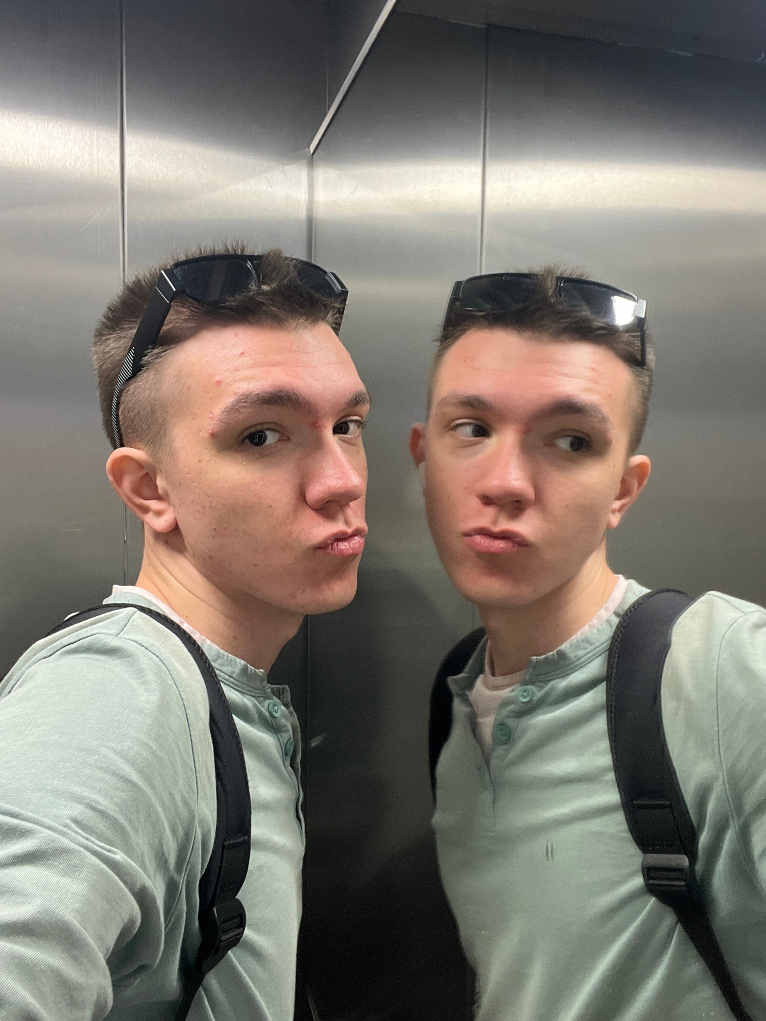 The image features a young man, viewed from the chest up, with his face reflected in a mirrored surface. He is wearing a light blue collared shirt with two buttons visible at the neckline and a black strap that appears to be part of a backpack or messenger bag. He has short, dark hair and is wearing dark-rimmed sunglasses pushed up onto his head. The man is looking directly at the camera, with a neutral expression. His skin has a few blemishes, and he appears to be indoors, as suggested by the smooth, metallic surface of the mirrored wall and the indoor lighting. The reflection is clear and shows his face and upper body accurately. The overall impression is a casual selfie taken in an elevator or a similar mirrored interior space. There is no visible text in the image. The location context of Romashkovo, Russia is provided but not visually evident in the image itself.