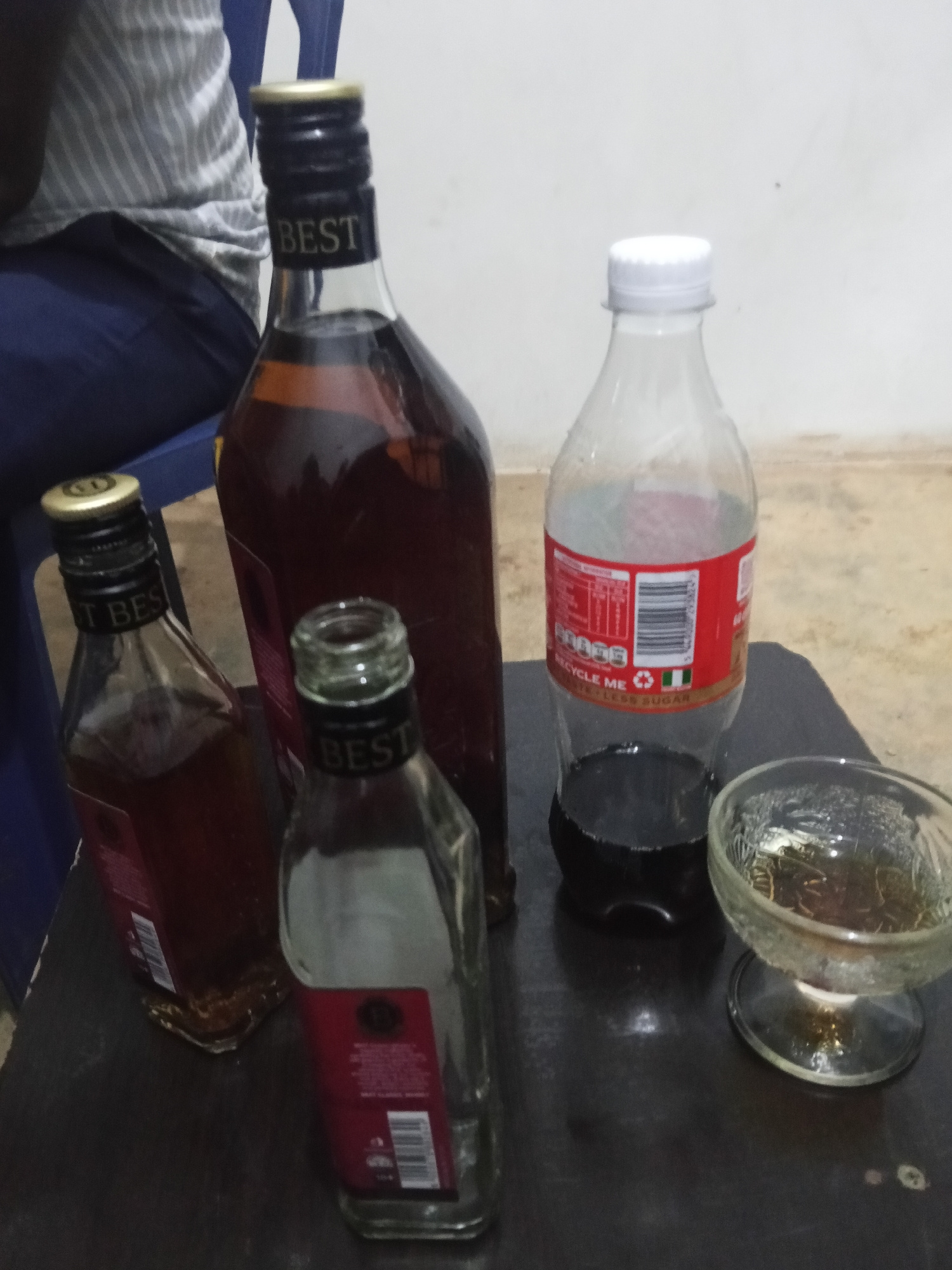 A medium shot, taken from a slightly elevated angle, shows a dark wooden surface covered with several bottles and a small glass dish. To the left, a tall, dark glass bottle with a gold cap and black label is partially visible. Next to it, a smaller, similar bottle is also present. In the center, a large, dark glass bottle with a gold cap is the most prominent object. To the right of this bottle, a clear plastic bottle with a white cap contains a dark liquid, likely soda, and has a red label with white text. In the foreground, a small, clear glass dish with a stem holds a dark amber liquid. The background shows a blurry image of a person sitting on a blue chair, wearing a striped shirt and dark pants. The overall scene appears to be an indoor setting with a light-colored wall in the background. The items suggest a setting where drinks are being consumed, possibly in Jos, Nigeria, given the detail provided.