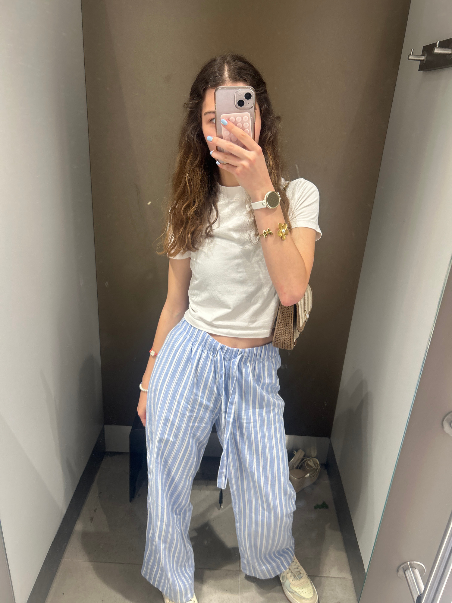 In what appears to be a fitting room, a young woman is captured taking a selfie in a mirror. She is wearing a white t-shirt and light blue and white striped pants. Accessories include a white watch on her left wrist and a gold bracelet with a flower design. She is also holding a smartphone with a pink case. The background is a neutral-toned wall, characteristic of a retail environment. The image does not provide any information that would allow for its localization to the city of Ruse, Bulgaria.