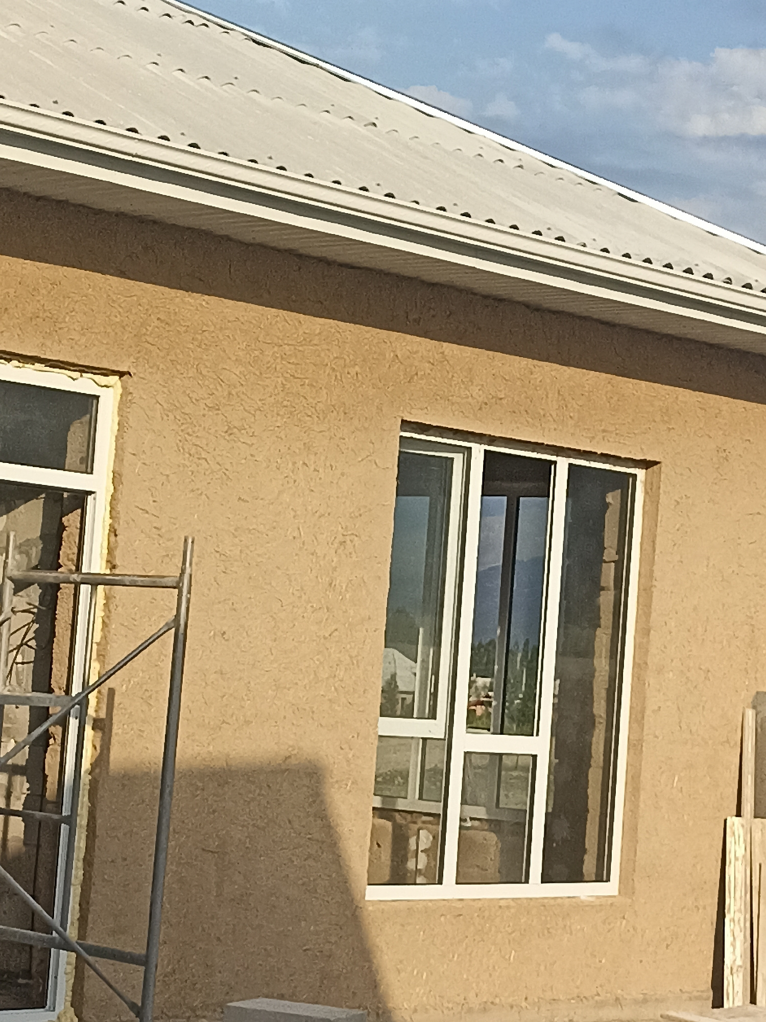 The image depicts the exterior of a building under construction in Кош-Дөбө, Kyrgyzstan. The building features a light-colored, textured stucco finish and a corrugated metal roof. Several windows with white frames are visible, some appearing to be in place while others might be awaiting installation. Scaffolding is present next to one of the windows, suggesting ongoing work. The environment appears to be an outdoor setting, likely a residential or commercial construction site, with hints of the sky and possibly surrounding structures visible through the windows. The overall scene conveys a sense of ongoing development and building activity.
