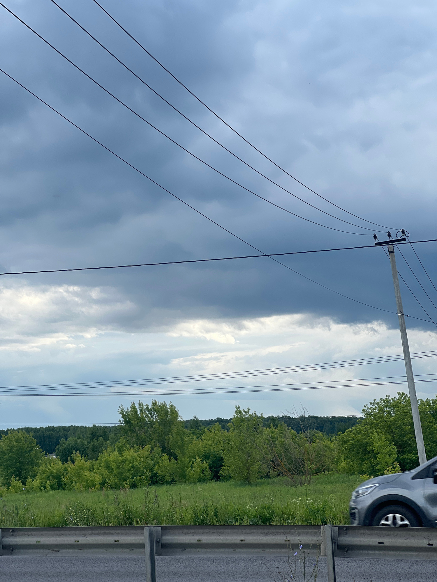 This is a vertically oriented, outdoor, eye-level medium shot of a roadside scene in Kosyakino, Russia. The dominant subjects are power lines and a telephone pole that cut across the frame diagonally from the lower left to the upper right. These lines are silhouetted against a sky that is half-covered by dark, ominous storm clouds on the left, transitioning to a brighter, partly cloudy sky on the right.
Below the sky, a dense, green forest lines the horizon. In the foreground, on the right side of the frame, a gray car is partially visible, parked on the shoulder of a road. A metal guardrail runs along the edge of the road, separating it from the grassy verge and the forest. The grass is speckled with small white flowers.
The overall impression is one of an approaching storm. The contrast between the dark clouds and the bright, sunny portions of the sky suggests a dynamic weather system. The presence of the car and guardrail indicates a rural or semi-rural roadside environment. The time of day is not definitively clear, but the lighting suggests it could be late afternoon or early evening. There is no visible text in the image.