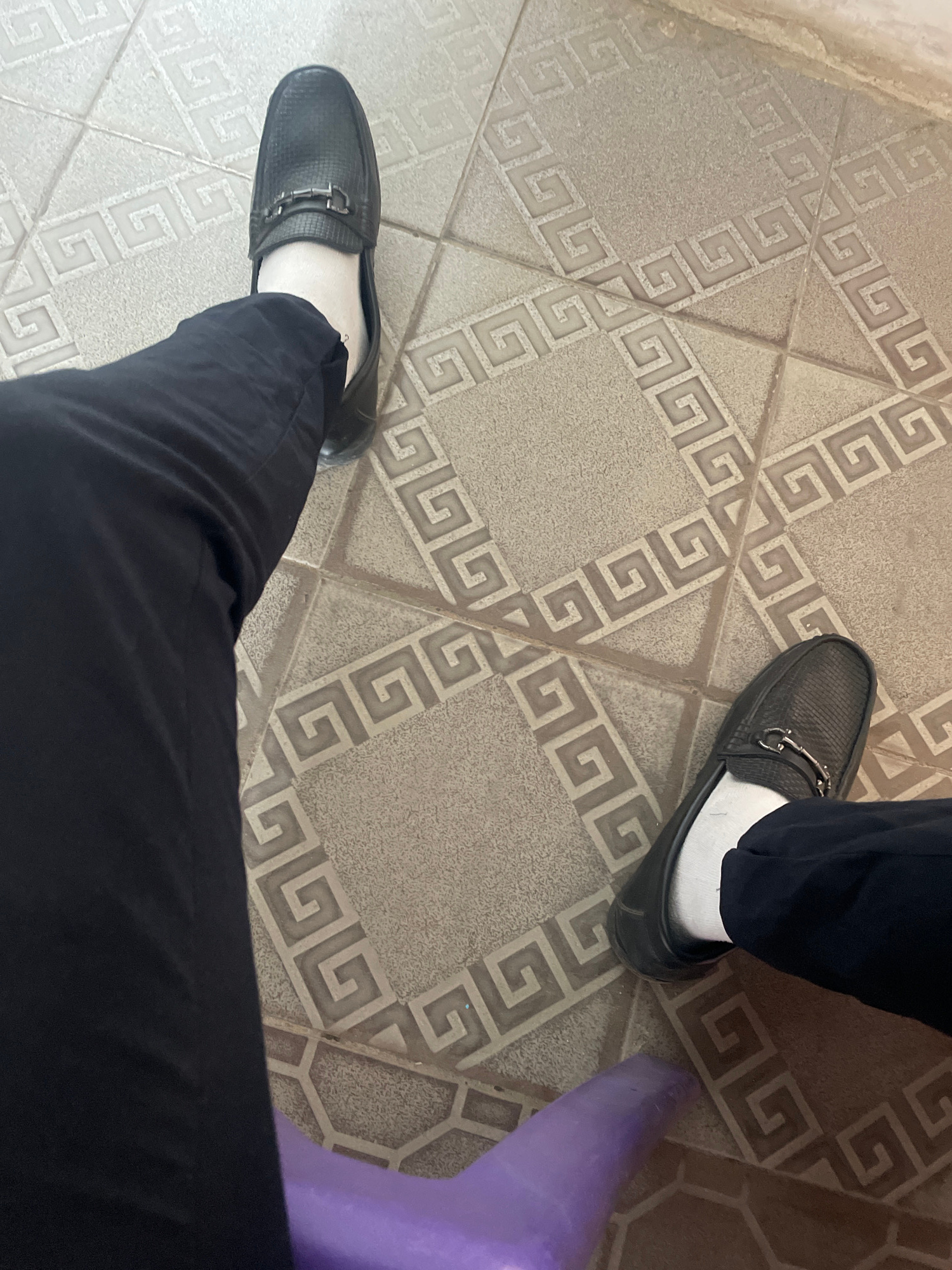 The image shows a close-up view of two people's legs and feet. They are both wearing black loafers and white socks, with black pants. The floor is tiled with a geometric pattern resembling a Greek key motif, in shades of beige and gray. To the right of the frame, a purple object, possibly a stool or chair, is partially visible. The lighting suggests it is daytime, with no indication of specific weather conditions. The scene appears to be indoors, possibly in a home or public space with tiled flooring. The focus is on the footwear and lower legs of the individuals, implying a casual or everyday setting. No text is visible in the image. The context provided indicates the location is Abuja, Nigeria.