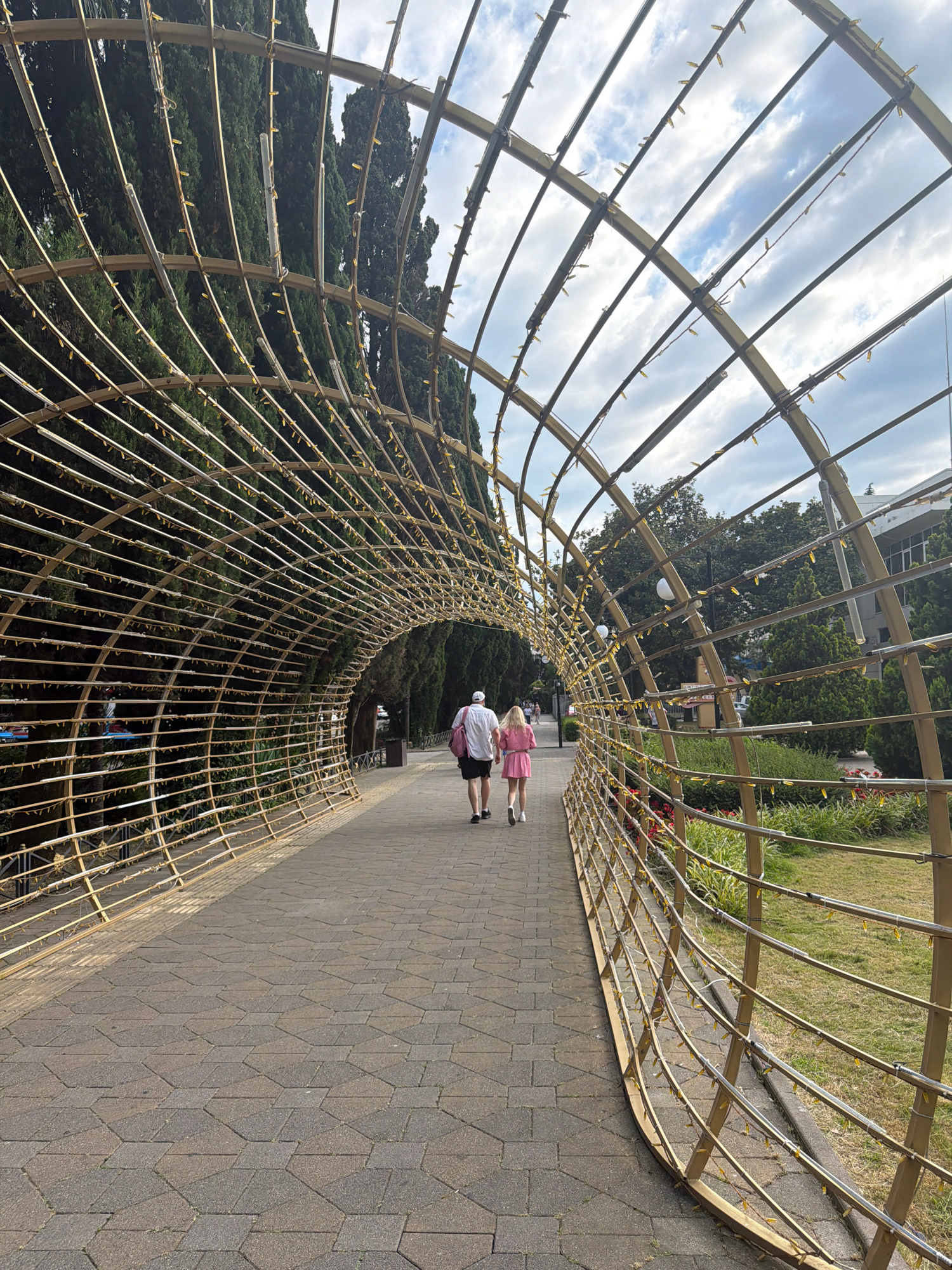 This image captures a view looking through a golden, arching tunnel structure adorned with small yellow lights. The tunnel is constructed of intersecting metal bars forming a lattice. On the left, tall, dark green trees line the pathway. On the right, a grassy area with some bushes and red flowers is visible, followed by buildings and more trees in the distance. A paved walkway runs through the center of the frame, bordered by the tunnel on the left and the grassy area on the right. Two individuals, a man and a woman, are walking away from the viewer down the pathway within the tunnel structure. The man is wearing a white t-shirt and black shorts, with a white cap. The woman is wearing a pink dress and carrying a pink bag over her shoulder. The sky above is partly cloudy. The scene is likely located in Sochi, Russia, given the context of similar architectural elements and vegetation often found in the city's parks and public spaces.