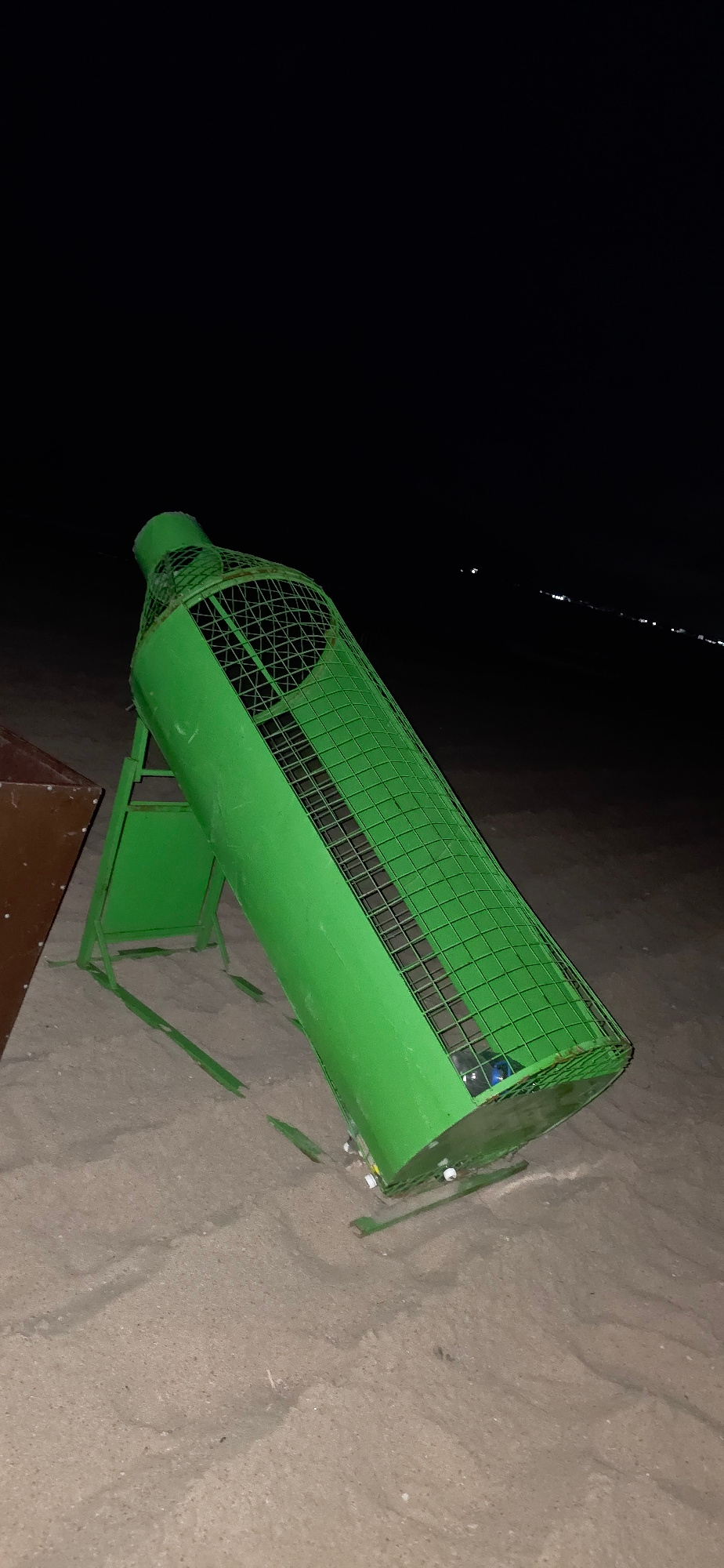 A bright green, cylindrical trash receptacle with a wire mesh front is positioned on a sandy surface. The receptacle is angled, with its wider end on the right and tapering towards the left. It is supported by a green metal stand. The background is a dark, almost black expanse, possibly representing water or a very dark sky, with some faint, shimmering reflections visible on its surface. The overall scene suggests an outdoor setting, potentially near a body of water at night or in low light conditions. There is no visible text in the image.