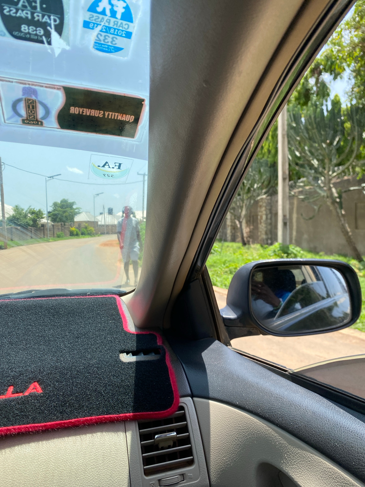 The image shows a view from inside a vehicle, looking through the windshield. In the foreground, a black car mat with red trim is visible. The windshield has several stickers, including one with the text "F.A." and what appears to be a logo. Outside the vehicle, a street is visible with houses and trees lining the sides. A person in a light-colored shirt and shorts is standing in the middle of the road. The scene appears to be in a residential area of Gura Top, Nigeria, judging by the architecture and vegetation. The sky is clear and the sun is shining. The side mirror of the car is also visible, reflecting part of the interior and the driver's hand.