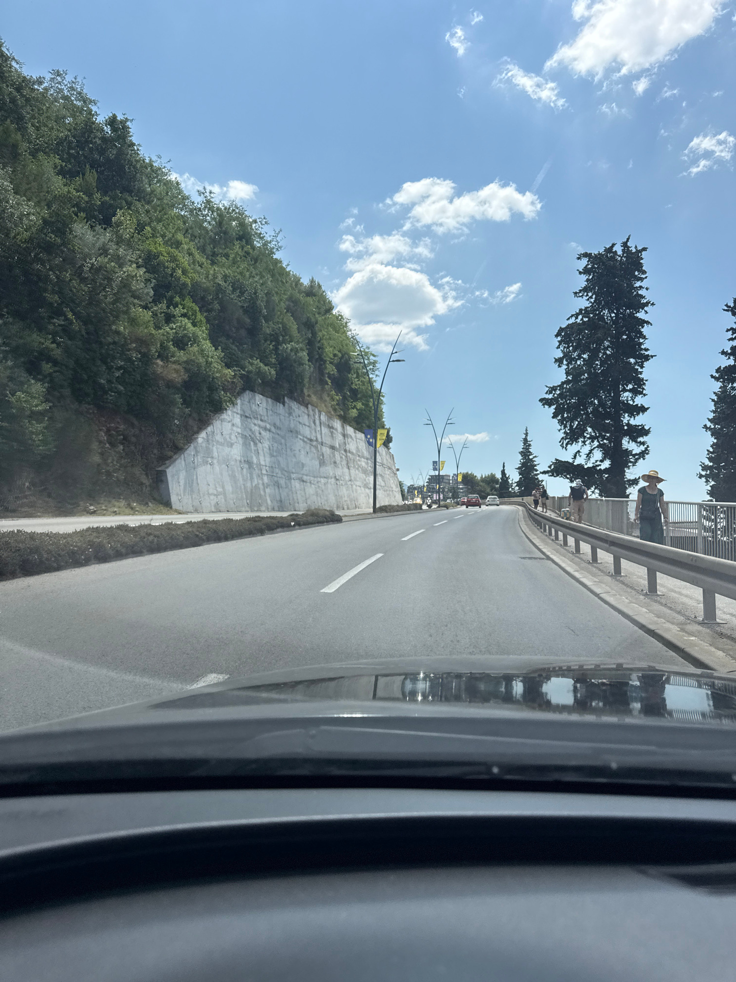 This is a first-person view from a vehicle driving on a road in Budva, Montenegro. The road is paved and has lane markings. To the left, a steep, tree-covered hillside rises, with a concrete retaining wall at its base. To the right, there is a sidewalk with a metal railing and several people walking. Tall coniferous trees are visible on the right side of the road. The sky is blue with scattered white clouds. There are other vehicles on the road in the distance. The foreground shows the dashboard and windshield of the vehicle.