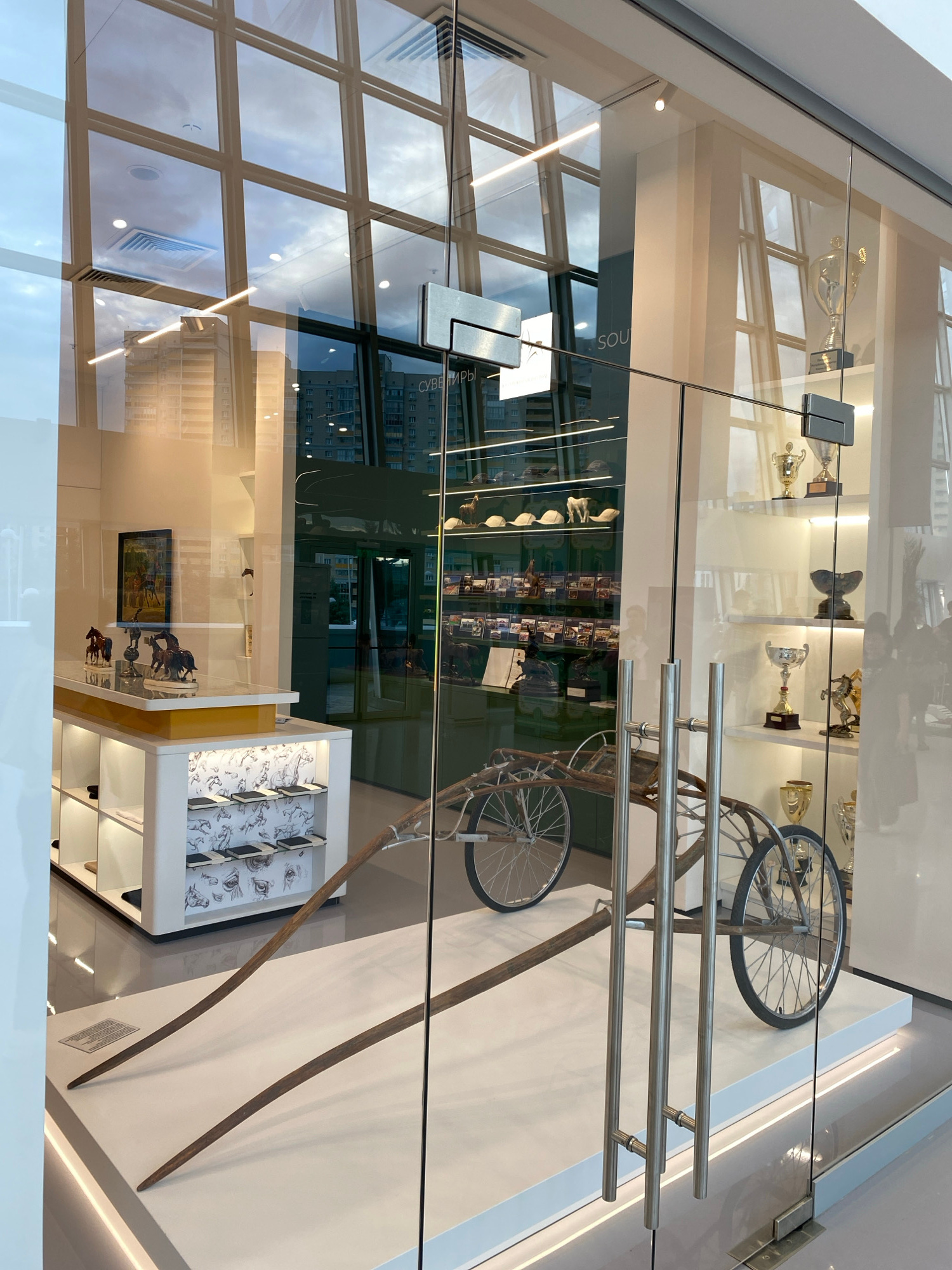 This image shows a storefront display window, possibly in Kazan, Russia, featuring a vintage horse-drawn sulky. The sulky is positioned centrally on a white platform, with its large spoked wheels and wooden shafts prominently displayed. The display is illuminated by a soft, warm light, creating reflections on the glass.
Inside the store, visible through the glass, is a collection of trophies displayed on shelves, suggesting a connection to racing or equestrian achievements. Various other items, including what appear to be figurines of horses and artwork depicting equestrian scenes, are also visible. The background reflects a modern building and the sky. The overall impression is that of a shop specializing in equestrian memorabilia or trophies.