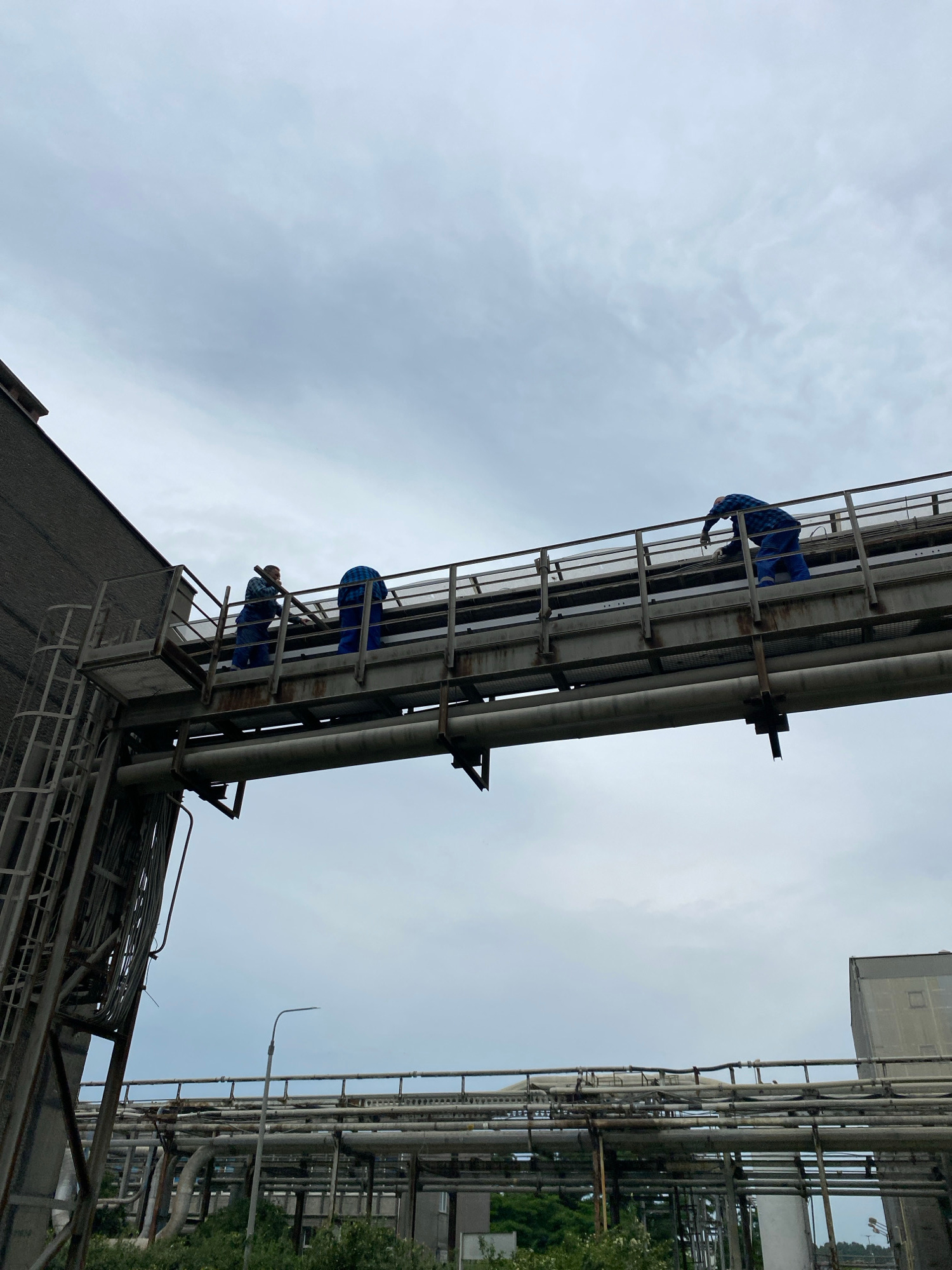 The image shows an industrial facility with several workers performing maintenance on elevated piping. In the foreground, a large, dark, textured building wall extends from the left edge of the frame upwards. Attached to this wall is a metal staircase and platform leading to an elevated walkway.
On the walkway, three individuals wearing blue jumpsuits are visible. The first worker, on the left, is positioned near the platform, facing away from the camera, holding what appears to be a pipe or bar. The second worker is further down the walkway, also facing away, seemingly engaged in work on the railing or piping. The third worker, on the far right of the walkway, is leaning over, possibly working on equipment.
The walkway itself is constructed of metal grating and supported by beams. Below the walkway, a large pipe runs horizontally. In the background, more industrial piping and structures are visible, along with some greenery and a streetlamp. The sky is overcast with clouds. The scene appears to be an outdoor industrial site, possibly a factory or refinery, under a cloudy sky. The location is not specified as being in the city of Police, Poland.
