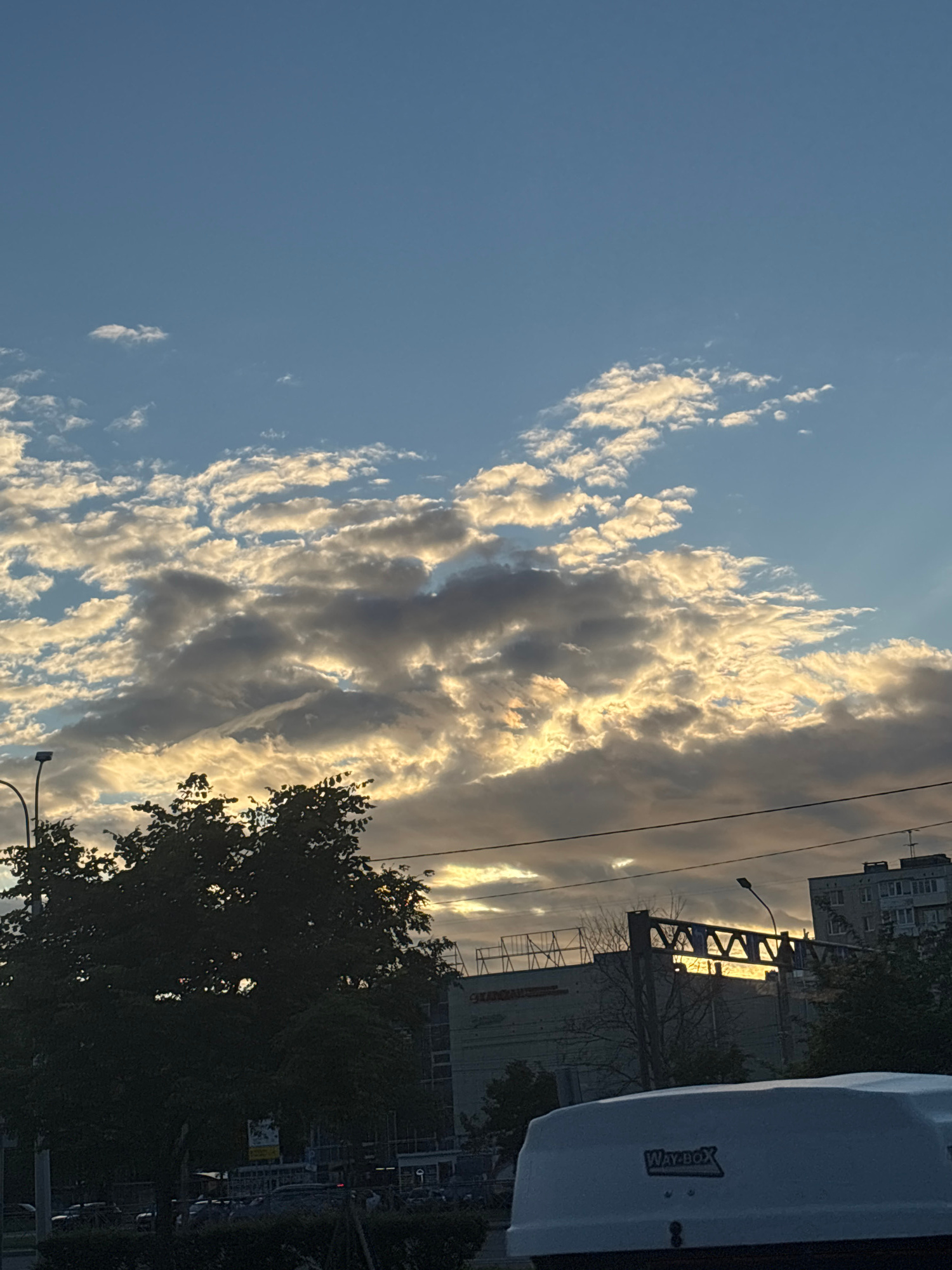 This media file displays a sky filled with dramatic, illuminated clouds against a clear blue sky. The sun is setting or rising, casting a golden light on the clouds, creating a contrast between the bright edges and darker, shadowed undersides. Below the sky, a silhouetted tree is visible on the right side, its leaves dark against the lighter sky.
Further down, hints of an urban setting emerge. There are buildings, one appearing to be a multi-story apartment or office building with visible windows. A large, white object, possibly a vehicle or storage container, is in the foreground on the right. It has "Way-Box" printed on its side. Power lines are visible crisscrossing the scene, and a street lamp can be seen. The overall impression is of an urban landscape at dusk or dawn, with the natural beauty of the sky taking prominence. The lighting suggests a late afternoon or early morning time of day.