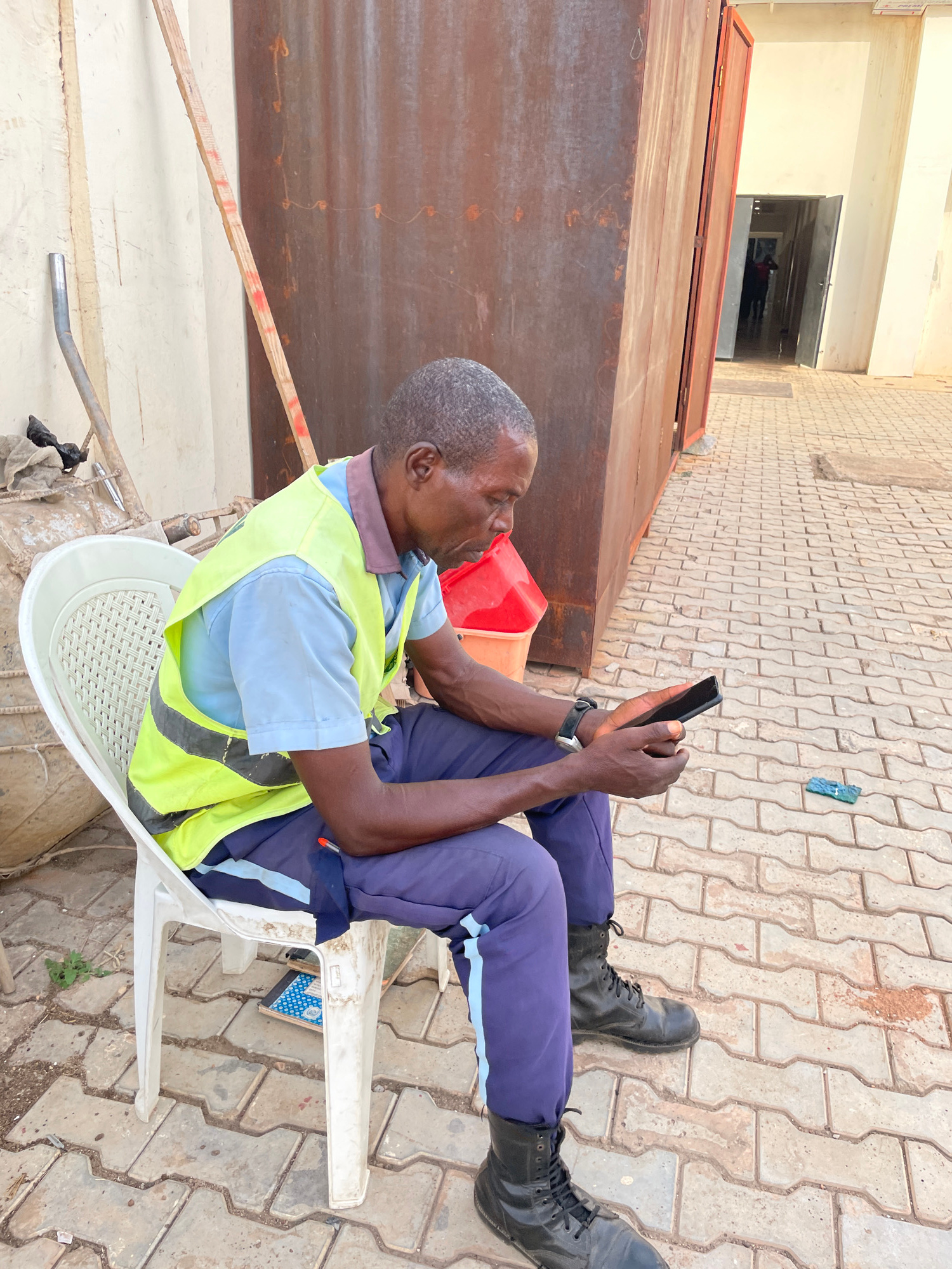 A middle-aged Black man sits on a white plastic chair, facing right, and looking down at a black smartphone he holds in his hands. He wears a blue short-sleeved shirt, a bright yellow safety vest with reflective gray stripes, dark blue pants with a light blue stripe down the side, and black combat boots. A red pen is tucked into his pants pocket on the right side. To his left, a red plastic bucket is visible. Behind him, a large, rusty metal structure forms a backdrop, with a lighter colored wall to the far left. The ground is paved with interlocking concrete bricks. In the distance, a doorway with an open double door leads into a dimly lit interior where a person is visible.