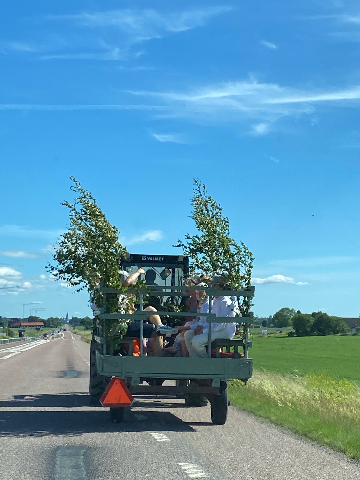 The image depicts a trailer, possibly from a tractor, being pulled along a road. The trailer is carrying several people, who appear to be young adults, some dressed in white shirts. They are seated on benches within the trailer. There are branches with green leaves adorned on the trailer and around the people, suggesting a festive occasion, perhaps a midsummer celebration given the location context of Uppsala, Sweden. The trailer has a "VALMET" logo visible on its side. A triangular orange reflector is attached to the rear of the trailer.
The scene is set outdoors on a sunny day, with a clear blue sky and scattered white clouds. The road is paved, and to the right of the road, there is a lush green field bordered by trees. In the distance, there are buildings and possibly a church spire, hinting at a rural or semi-rural setting. The overall impression is one of a joyous and casual gathering, enjoying the summer weather and participating in a local tradition. The image is oriented vertically, with the sky occupying the top half and the road and trailer in the bottom half.