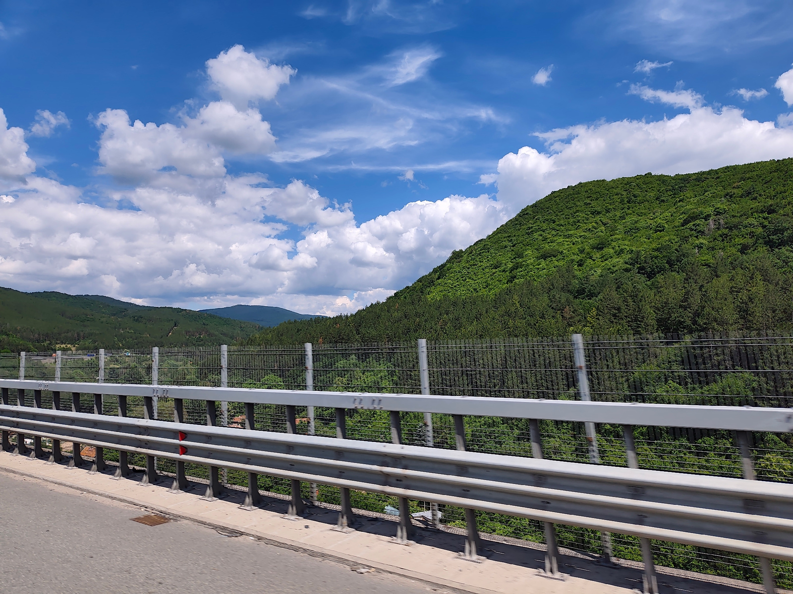 This image captures a scenic view from a bridge overlooking a lush, green forested mountain range under a bright blue sky filled with scattered white clouds. The foreground features the concrete and metal structure of the bridge, with its guardrails clearly visible. The bridge appears to be a highway overpass, with a gray asphalt road in the lower left corner. Behind the bridge, a metal fence separates the roadway from the descending slope, which is densely covered in vibrant green trees.
The mountain landscape extends into the distance, with rolling hills and more trees creating a sense of depth. The sky is a dominant element, with dramatic cumulus clouds adding texture and visual interest. The sun is high, suggesting it is daytime, likely mid-morning or early afternoon. The overall atmosphere is serene and picturesque, conveying a feeling of natural beauty and open space. No people or specific activities are depicted, focusing solely on the landscape and the man-made structure of the bridge. The location context is Potop, Bulgaria.