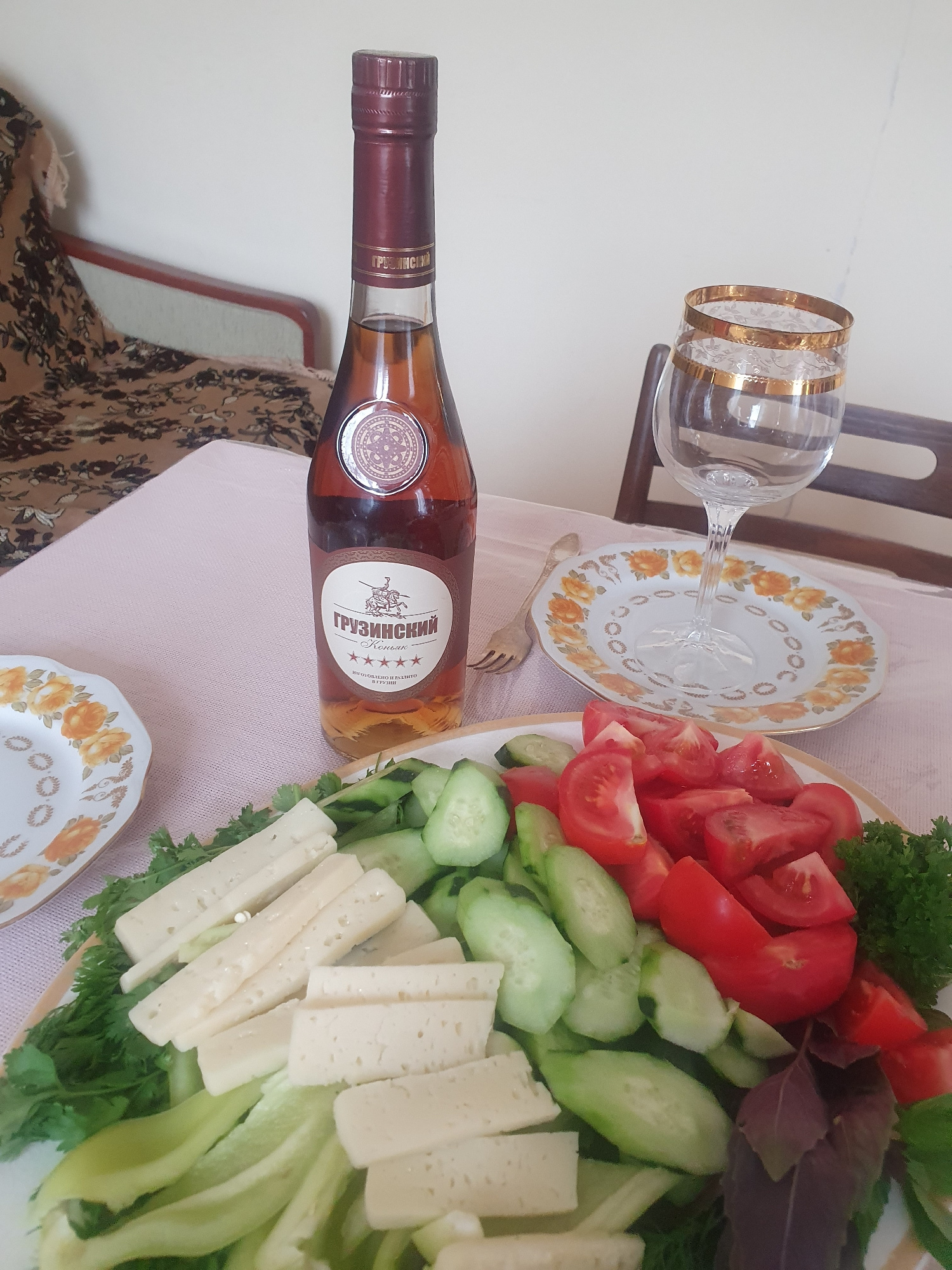 This is a high-angle, eye-level, medium shot of a table setting featuring a bottle of Georgian brandy, a glass, and a platter of food. The bottle of "Grozinsky" brandy is centrally located, with a prominent label depicting a knight on horseback. To the right of the bottle, an empty stemmed wine glass with gold rimming sits on a small decorative plate. In the foreground, a platter displays sliced white cheese, cucumber slices, chopped tomatoes, and parsley. The table is covered with a light pink tablecloth. In the background, a portion of a wooden chair and a patterned fabric are visible. The scene appears to be indoors, likely in a dining area, but its specific location in Almaty, Kazakhstan, cannot be determined from the visual information provided.