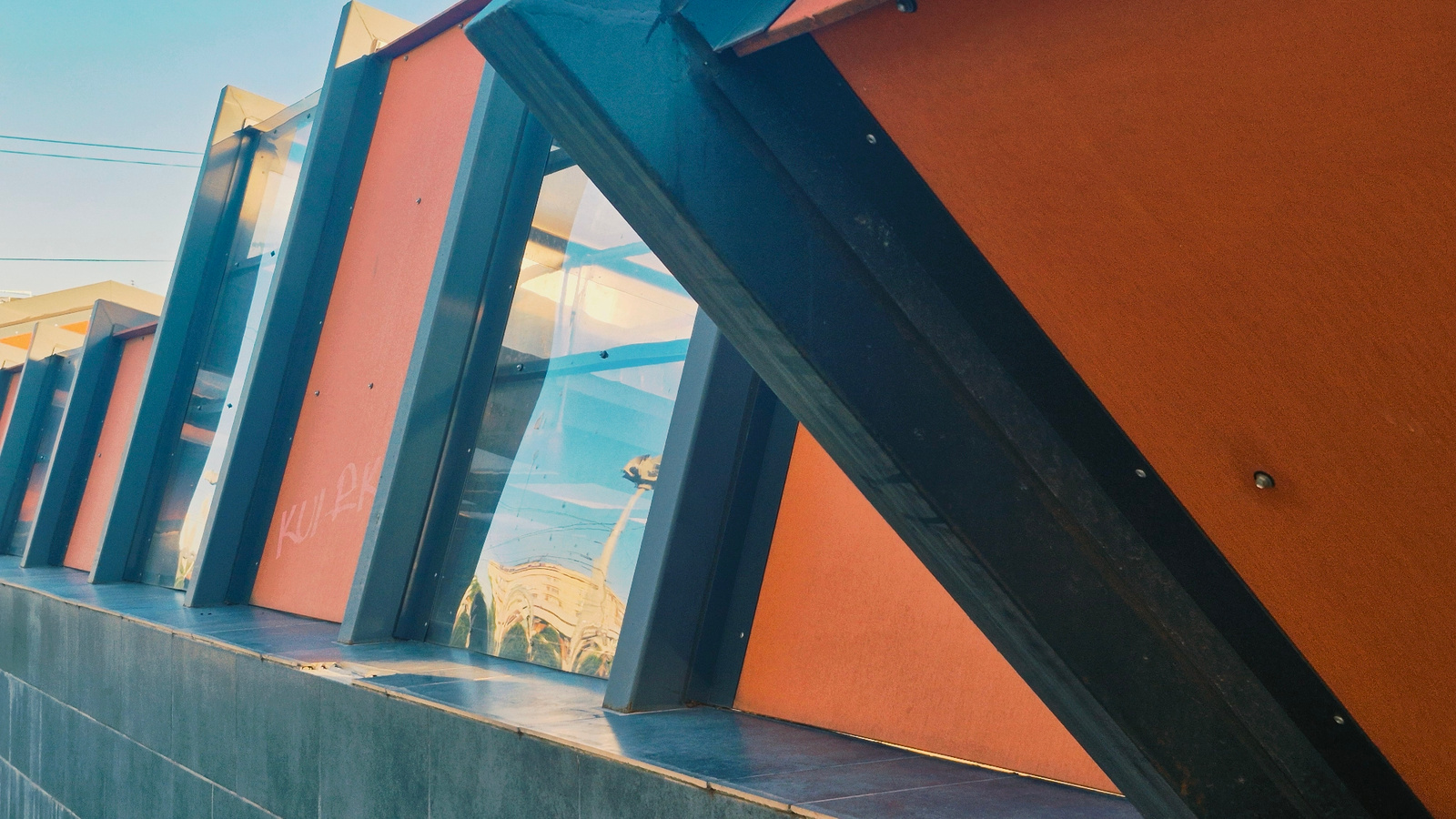The image depicts a section of an outdoor structure with a modern, angular design. The structure features a repeating pattern of dark metal beams angled inwards, framing panels of orange material and transparent glass. The glass panels reflect a distorted image of buildings and the sky, suggesting an urban environment. The orange panels have some light graffiti, appearing to be written in pink or red. The base of the structure is a textured gray wall. The overall lighting suggests it is daytime, with the sun casting shadows and highlighting the reflections. The scene is static and no people are visible. Based on architectural style and the presence of graffiti, this is likely a public space or an artistic installation.
The location in Chișinău, Moldova is the Children's Art School "Alexei Staveri" (formerly the Palace of Children and Youth). The specific architectural element shown is a decorative facade and canopy on the western side of the building.