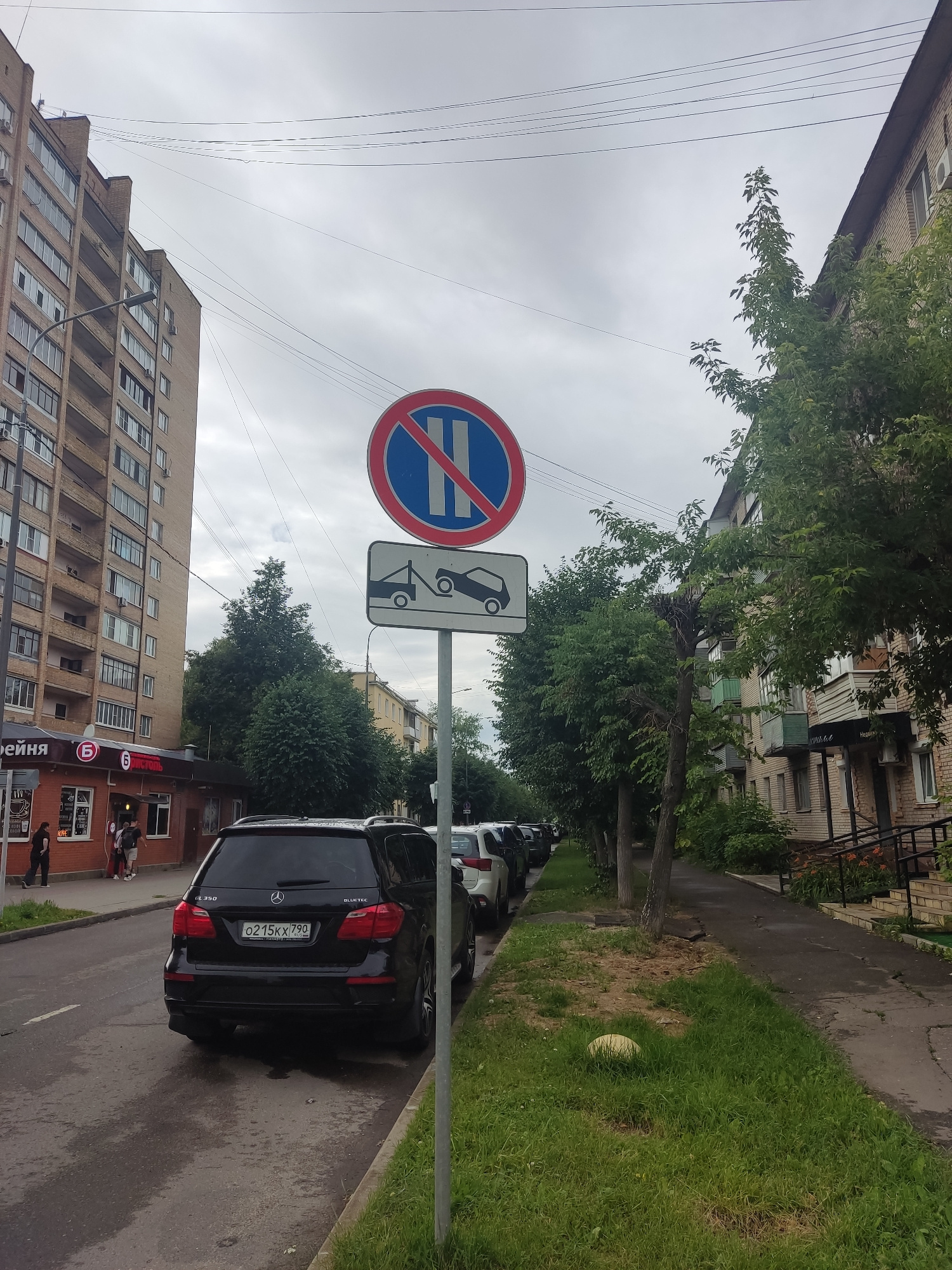 A street scene in Serpukhov, Russia features a two-story paved road with parked vehicles and a sidewalk lined with trees. In the foreground, a black Mercedes-Benz GL350 SUV is parked on the right side of the road, its license plate reading "o215kx790". Further down the street, several other cars are parked.
On the right side of the road, a sidewalk runs alongside buildings. A lamppost stands next to the sidewalk. Traffic signs are visible on a pole. The top sign is a blue circle with a red border and a red diagonal bar, indicating "No stopping". Below it, another sign shows a tow truck towing a car, signifying "Towing enforced".
The buildings on the left are tall, multi-story apartment blocks constructed with brick. On the right, there are lower-rise buildings with brick facades. The sky is overcast with grey clouds. The overall environment suggests an urban residential area on a cloudy day. A few individuals are visible on the sidewalk in front of a building on the left, which appears to be a small shop or cafe.