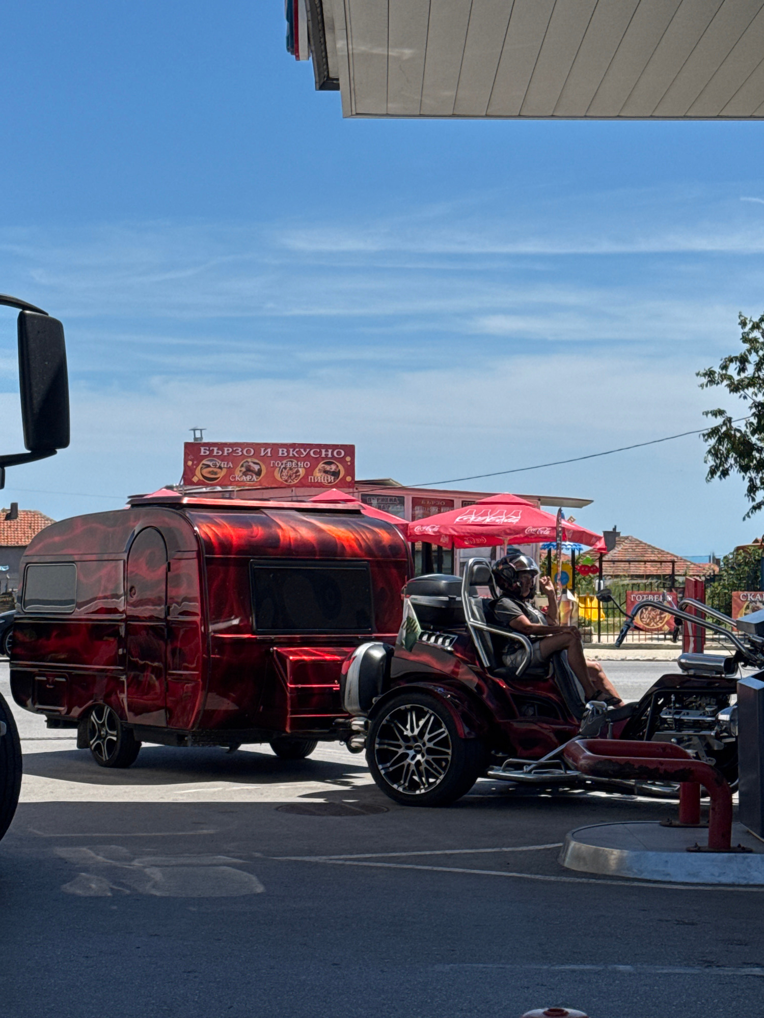 An eye-level medium shot captures a custom red and black flame-painted trailer being pulled by a motorcycle trike with a person wearing a helmet seated in the driver's position. The trailer features a rounded, retro design with two windows and a hinged door. Above the trailer, a sign reads "БЪРЗО И ВКУСНО" (Fast and Delicious) in Bulgarian, with smaller text indicating various food items like "СУПА" (Soup), "СКАРА" (Grill), "ГОТВЕНО" (Cooked), and "ПИЦИ" (Pizzas). Red umbrellas with Coca-Cola branding are visible in the background, along with buildings and trees under a clear blue sky with wispy clouds. The scene appears to be on a street in Byala, Bulgaria, possibly near a roadside eatery or a rest stop. The motorcycle trike has a chrome exhaust system and a distinctive wheel design. The person in the trike is looking towards the camera, with their left hand raised slightly.