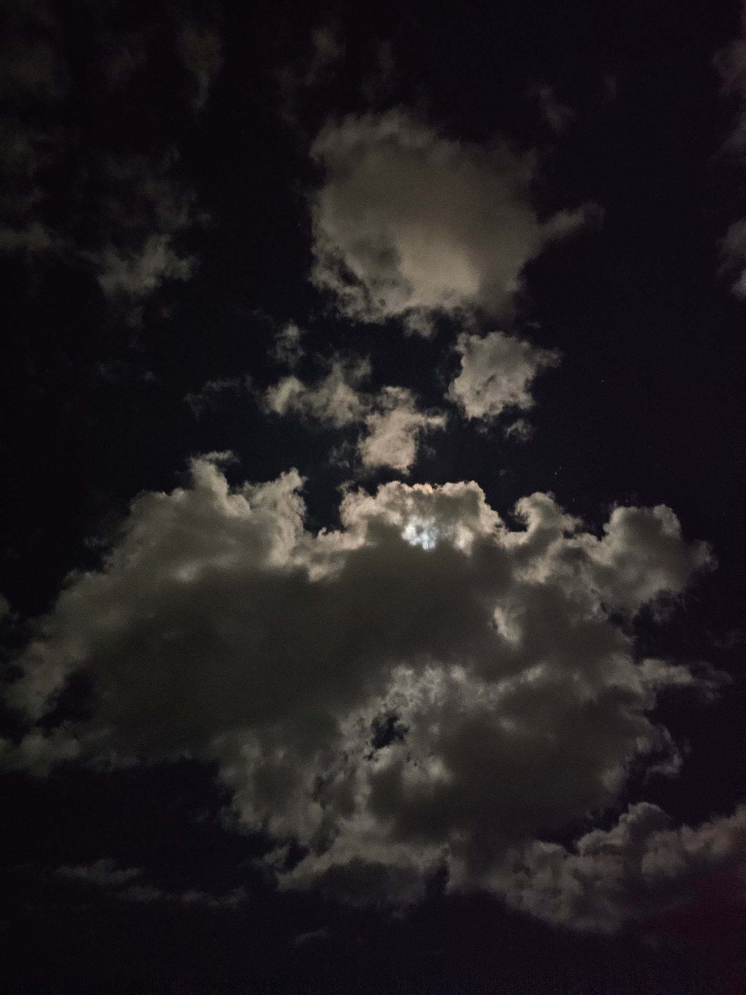 This is a photograph taken at night, likely in Denpasar, Indonesia, featuring a dark sky filled with clouds and the moon peeking through. The primary subjects are the clouds, which vary in density and luminosity. Some are thick and dark, obscuring the sky, while others are thin and wispy, catching the moonlight and appearing brighter. The moon itself is partially visible, its light illuminating the edges and undersides of the clouds.
The scene is a natural one, depicting the night sky. There are no people or man-made objects clearly visible. The activity, if any, is passive; it's a snapshot of the celestial landscape at a particular moment. The notable details include the time of day, which is clearly night due to the darkness of the sky and the presence of the moon. The weather appears to be cloudy, with intermittent breaks allowing the moonlight to shine through. There are no specific location cues beyond the general context of Denpasar, Indonesia. The image evokes a sense of quiet contemplation or perhaps a touch of mystery, given the dramatic interplay of light and shadow amongst the clouds. The lack of clear features and the atmospheric conditions suggest a serene or perhaps slightly foreboding mood. There is no visible text in the image.